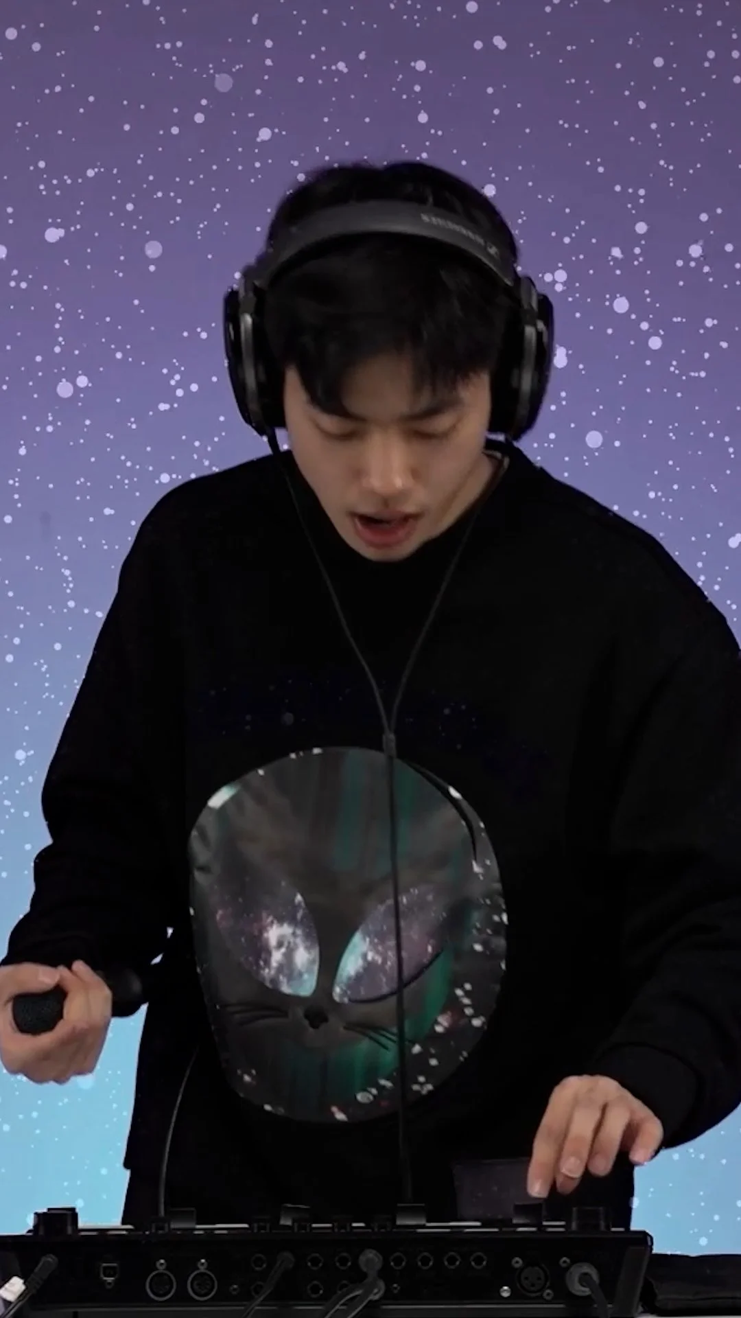 A DJ wearing headphones and a black sweatshirt with an alien face galaxy print, performing with DJ equipment against a starry purple background.
