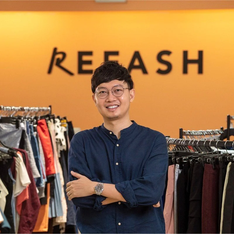 A man with glasses smiling and standing with his arms crossed in a clothing store, with the word REASH on the orange wall behind him.