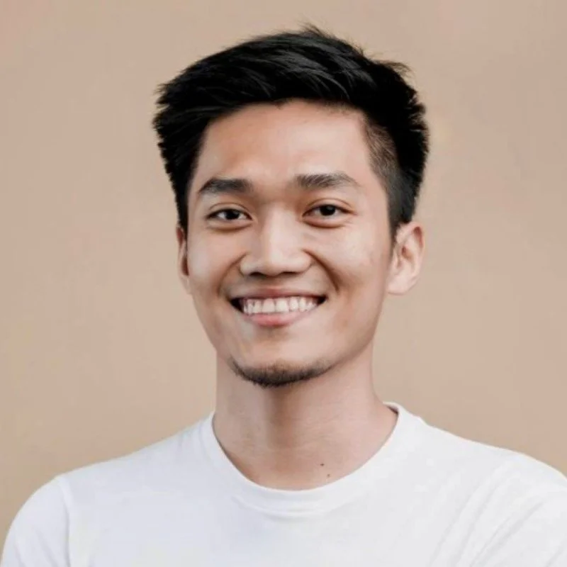 Portrait of a young man with dark hair smiling, wearing a white t-shirt against a beige background.