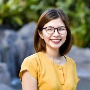 A young woman with shoulder-length brown hair wearing glasses and a yellow top, smiling outdoors with plants and rocks in the background.