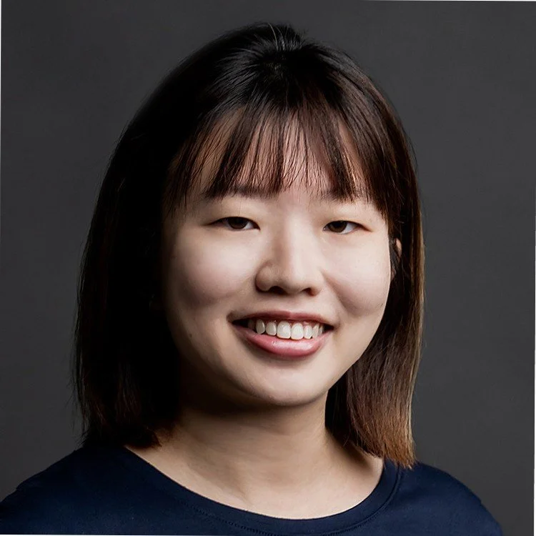 A portrait of a young woman with shoulder-length dark hair and bangs, smiling against a dark background.