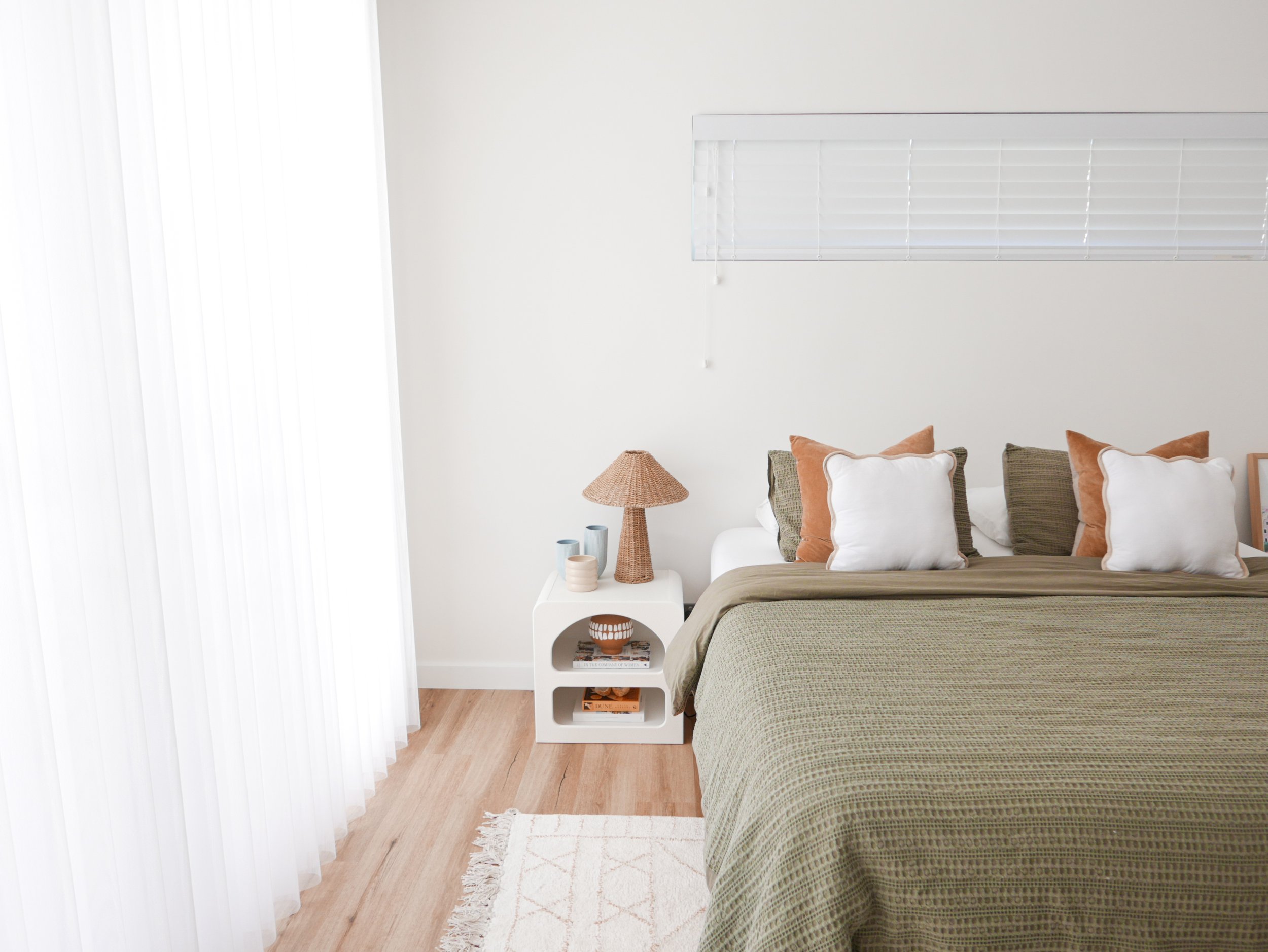 Calm and neutral styled bedroom with olive green waffle linen bedding, terracotta cushions, rattan lamp, and white arched bedside table on light timber floors — e-styling and Airbnb bedroom design by The Home Studio Co