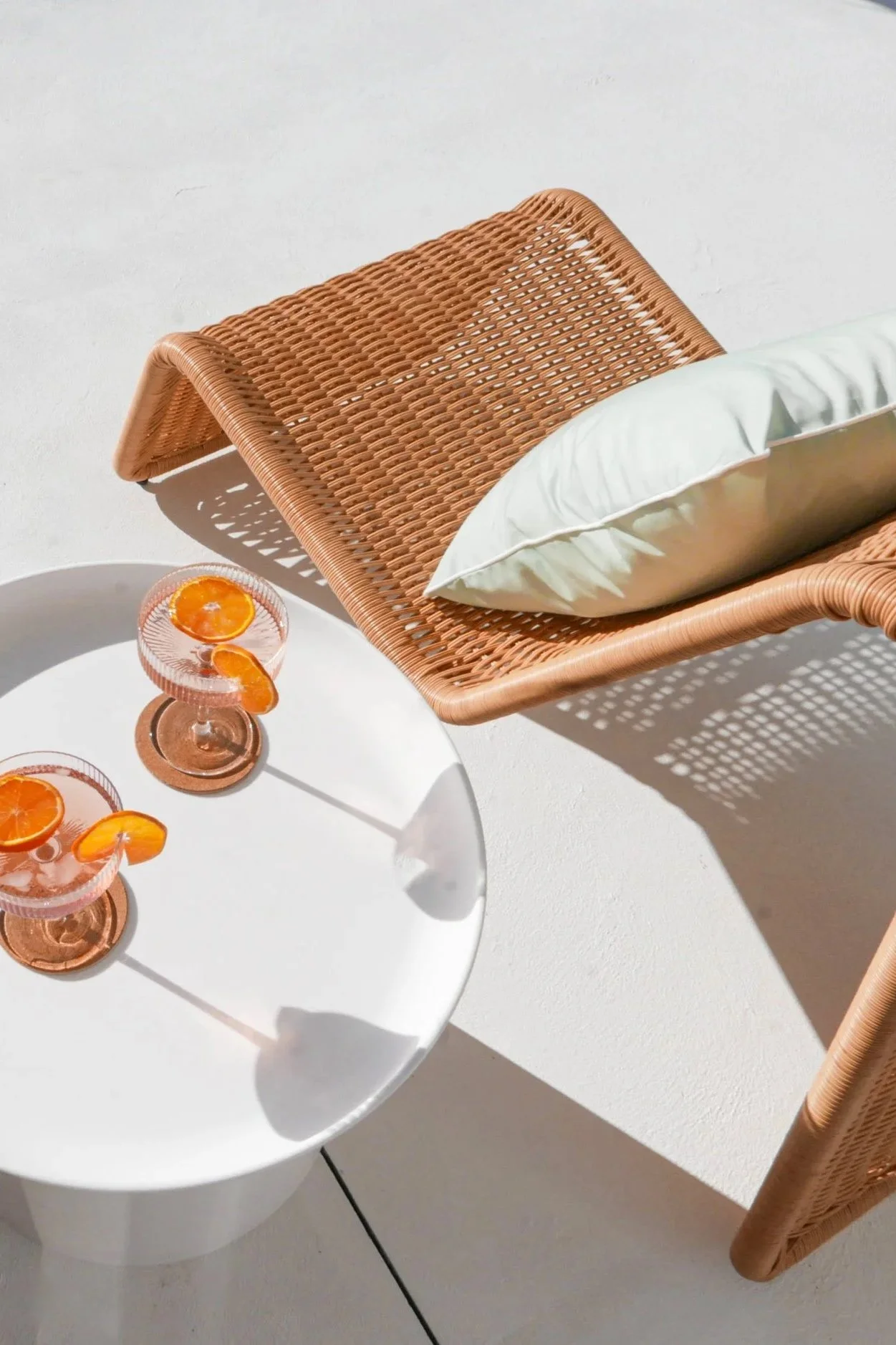 outdoor entertaining vignette with a tan woven rattan sun lounger, sage green cushion, white round side table, and two pink ribbed coupe glasses with orange slices in bright natural sunlight — Airbnb outdoor styling by The Home Studio Co