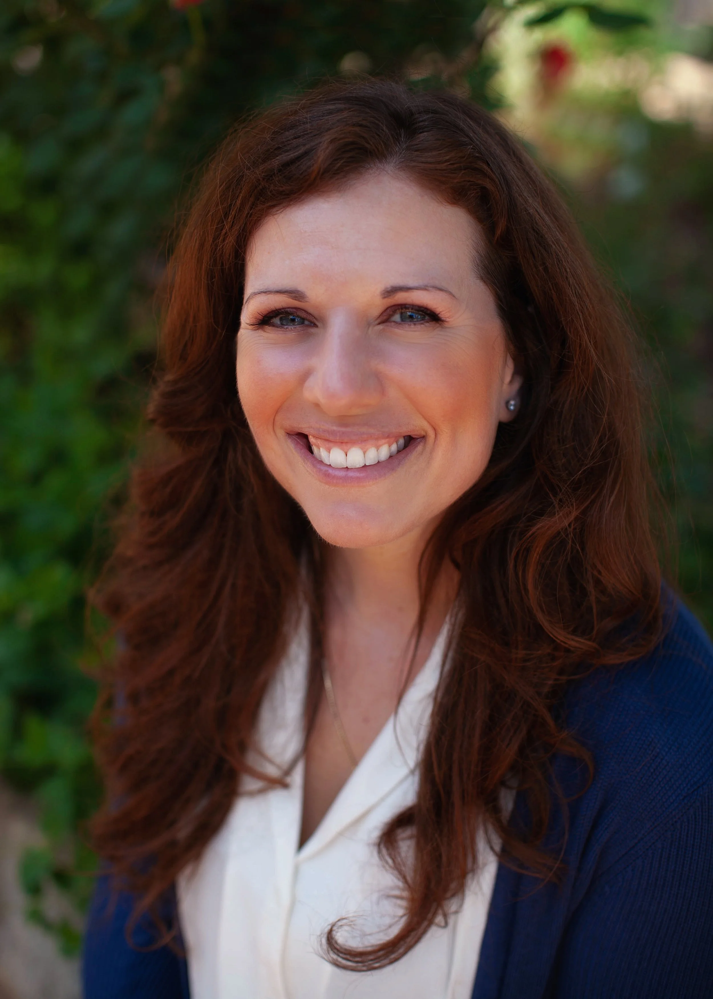 Smiling woman with long, wavy brown hair, wearing earrings, outdoors in a natural setting with greenery in the background.