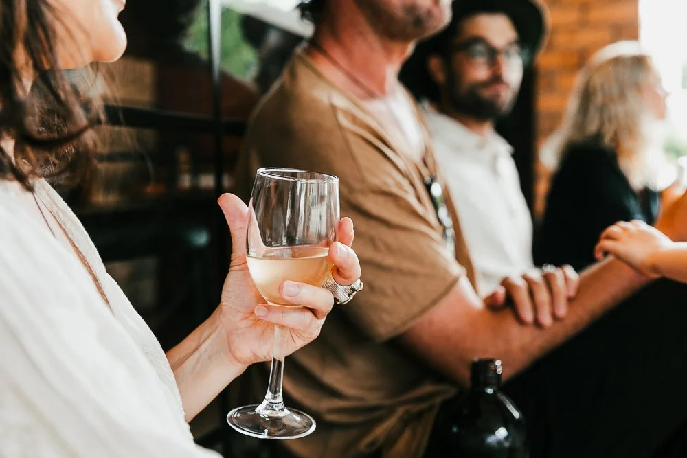 Natural Wine Shop & Bar: Person holding a glass of white wine in a social setting with others in the background.