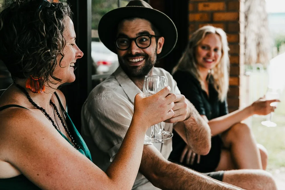 Natural Wine Shop & Bar: Three people sitting together, smiling, at a social gathering. The woman on the left is holding a glass, the man in the middle is wearing a hat and glasses, and the woman on the right is also holding a glass.
