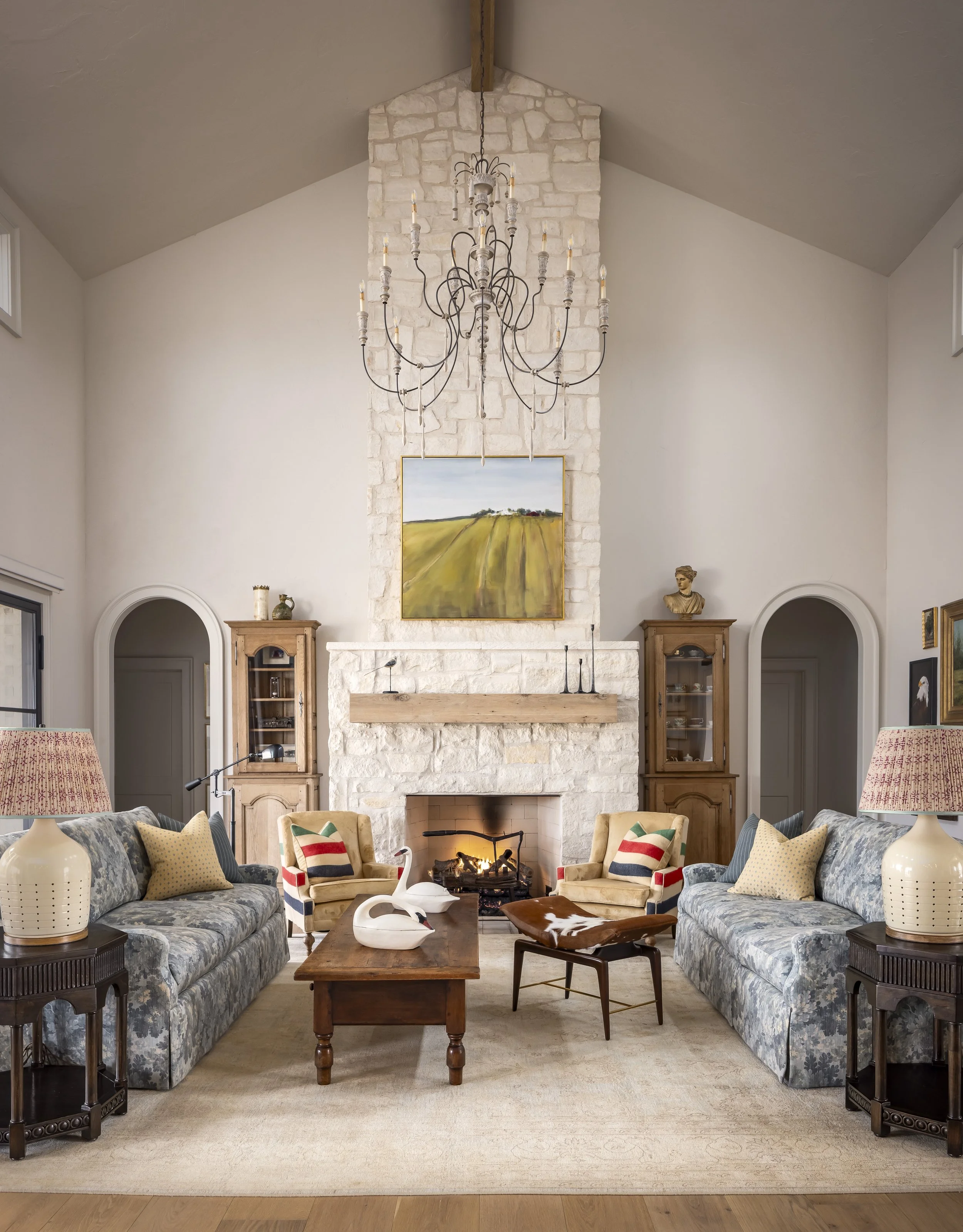 Elegant living room interior with stone fireplace, large chandelier, and artwork above the mantel. Symmetrical sofas and cozy armchairs surround a wooden coffee table. Decor includes swan sculptures, patterned cushions, and side tables with lamps.