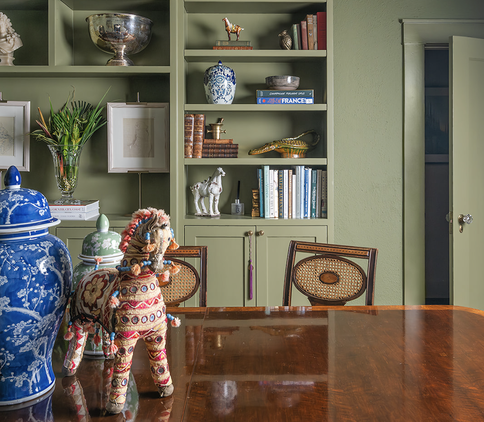 A dining room with a polished wooden table, green shelves filled with decorative objects and books, and a green wall. There are two chairs with woven backs, a large blue and white porcelain jar, a plush horse toy, and a glass vase with green plants on the table.
