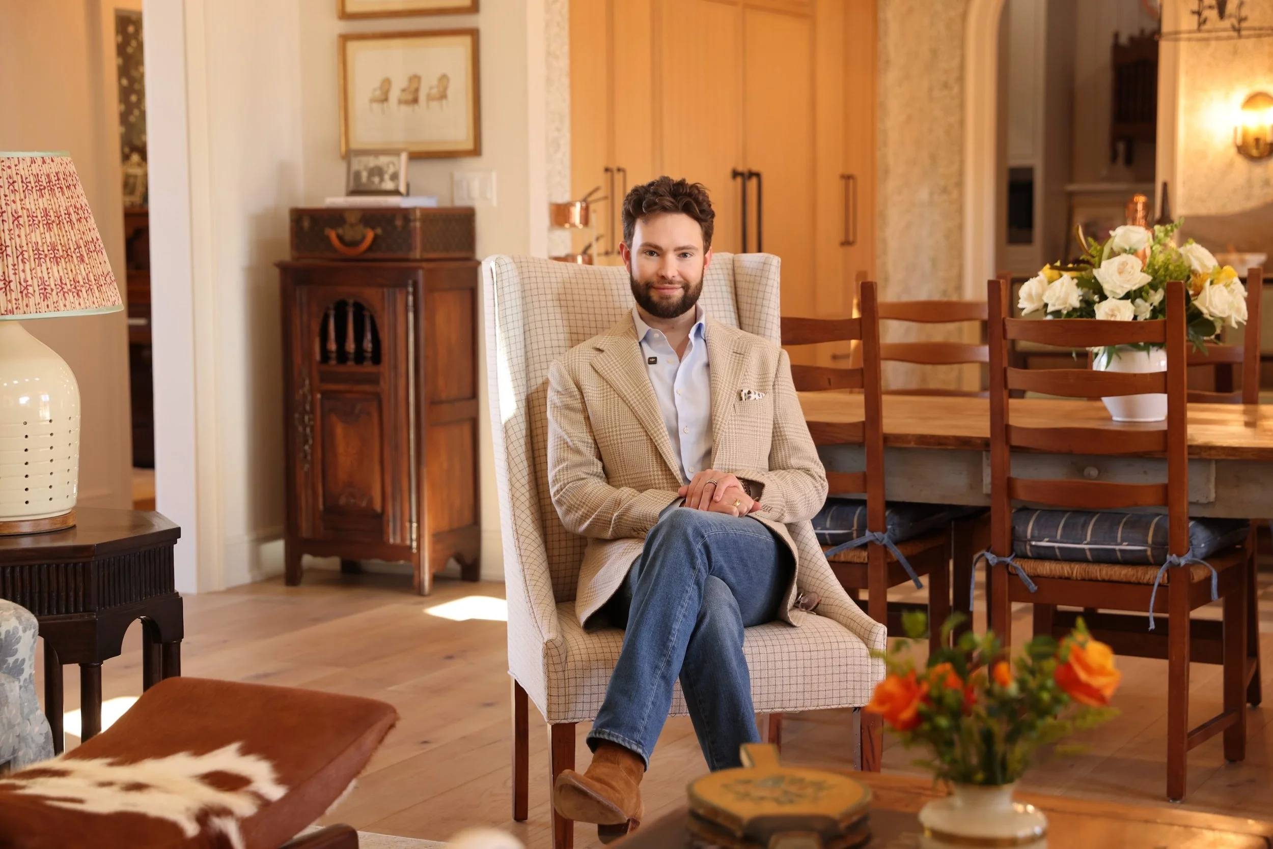 Fort Worth, Texas, Shayne Morrissey, interior designer, sitting in an armchair in a well-decorated living room, wearing a beige blazer and jeans. The room features a wooden dining table with chairs, a vase of flowers, a side table with a lamp