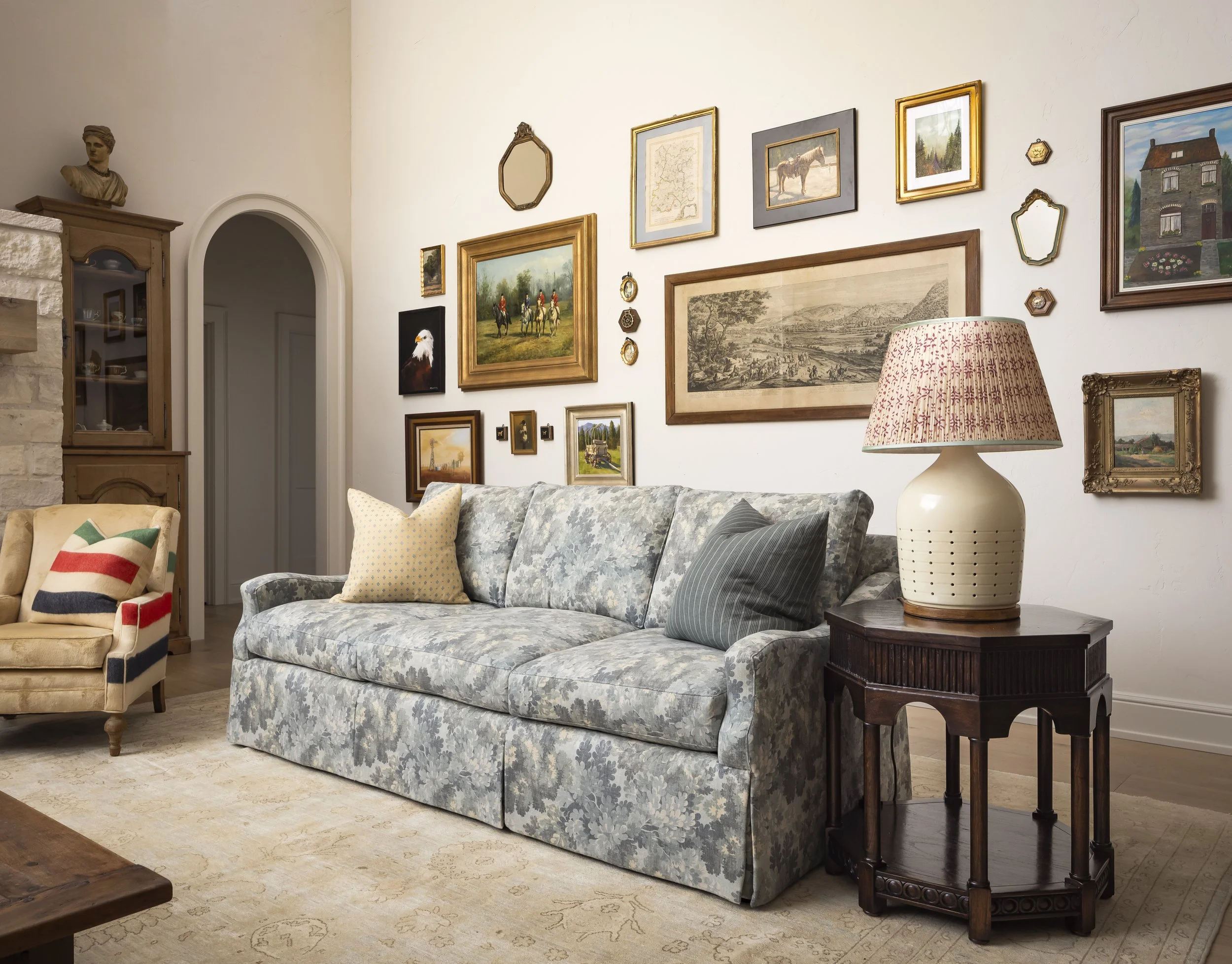 Vintage living room with a blue floral sofa, patterned armchair, and side table featuring a decorative lamp. The wall is adorned with various framed artworks and a sculpture above a cabinet.