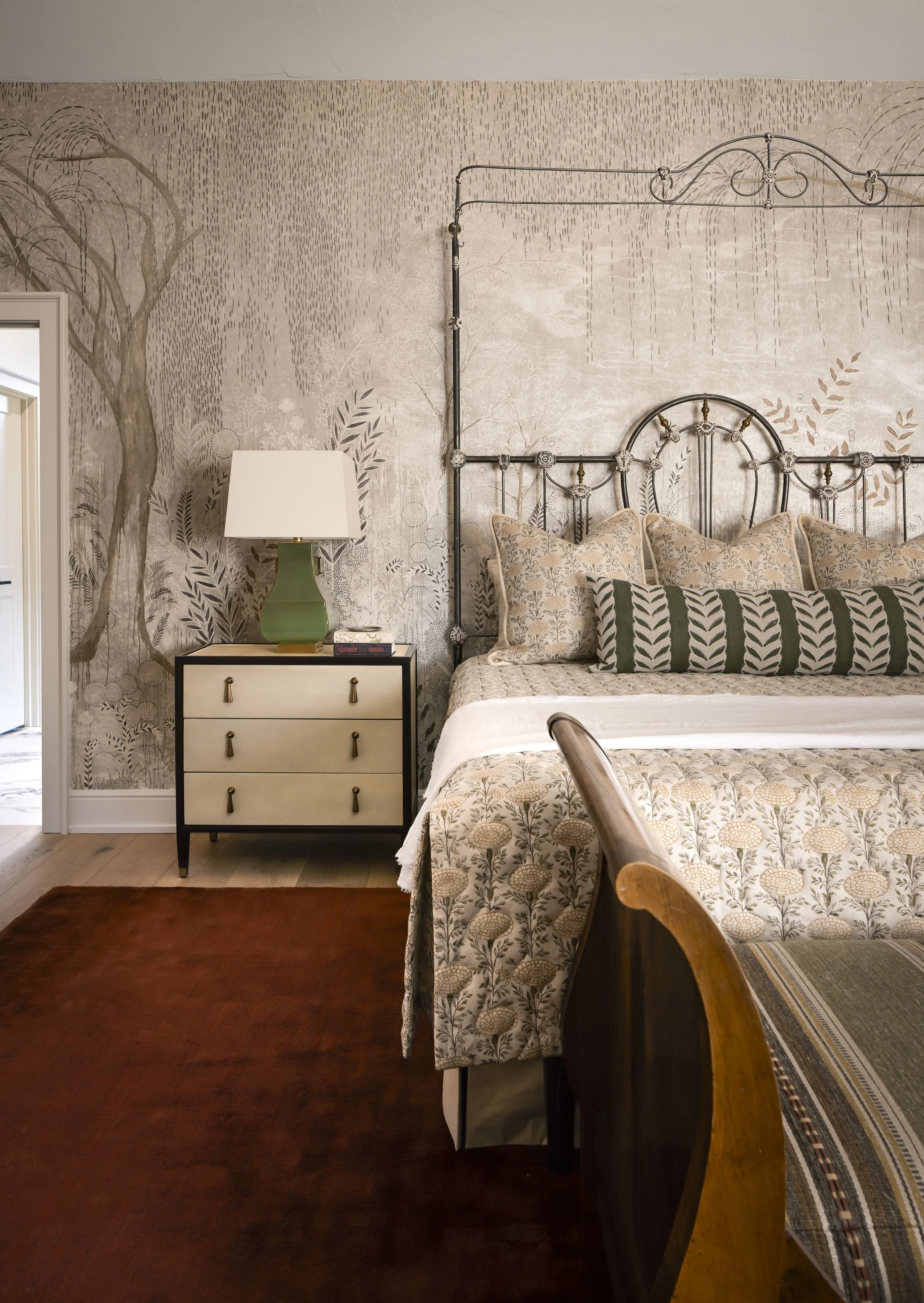 Cozy bedroom with vintage-style iron bed, floral bedding, green lamp on nightstand, and artistic wall mural.