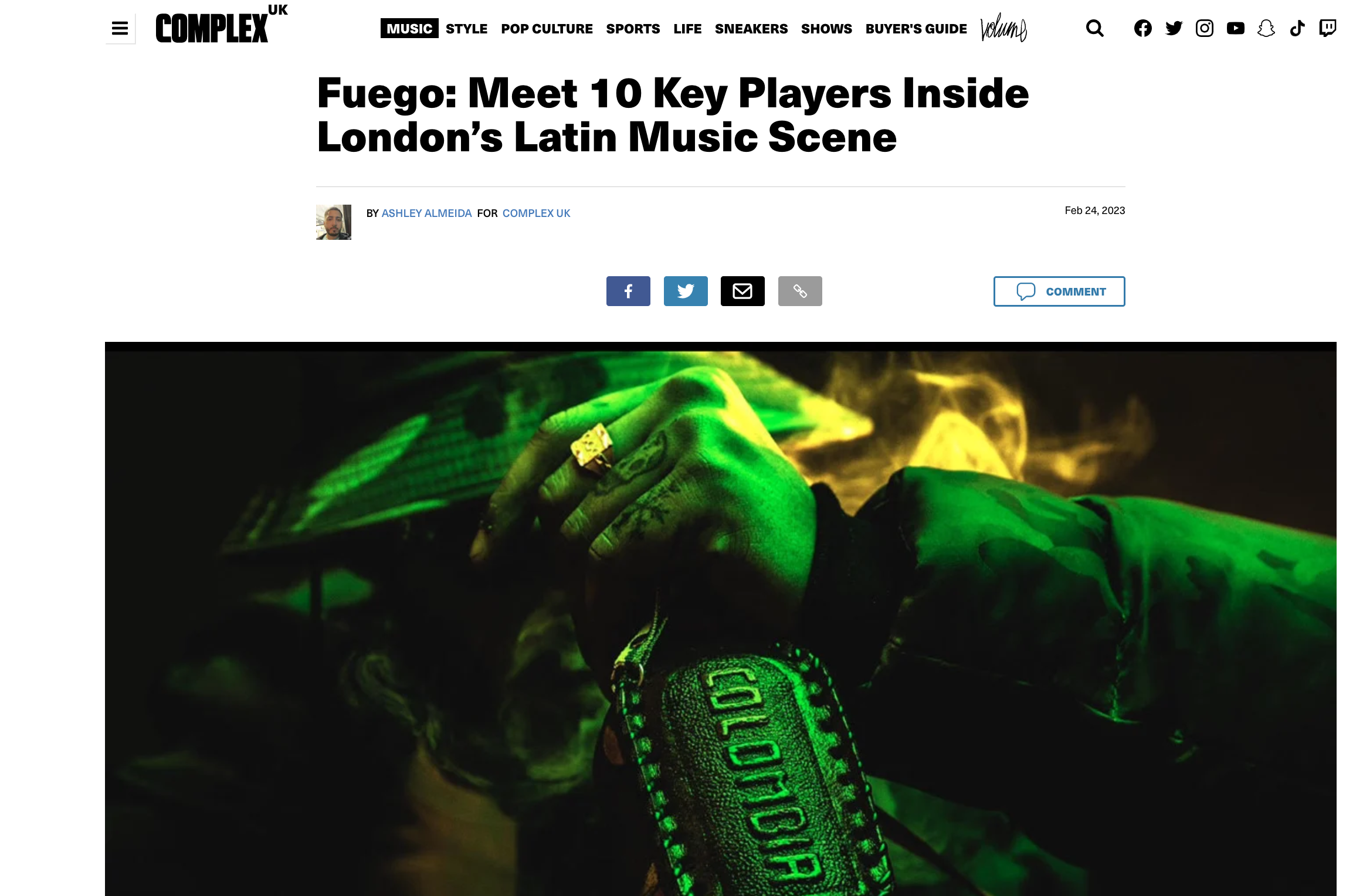 A person wearing a green digital camouflage jacket and a wristwatch with a large ring on their finger, resting their hands on a laptop, illuminated by green and yellow lighting.