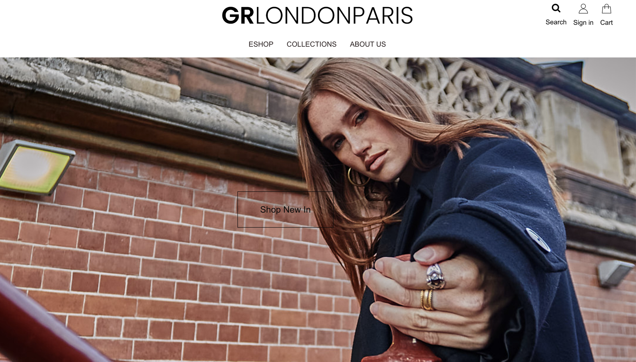 A young woman with long brown hair gazes into the camera while leaning on a red railing outside a brick building, wearing a navy blue jacket with rolled-up sleeves.