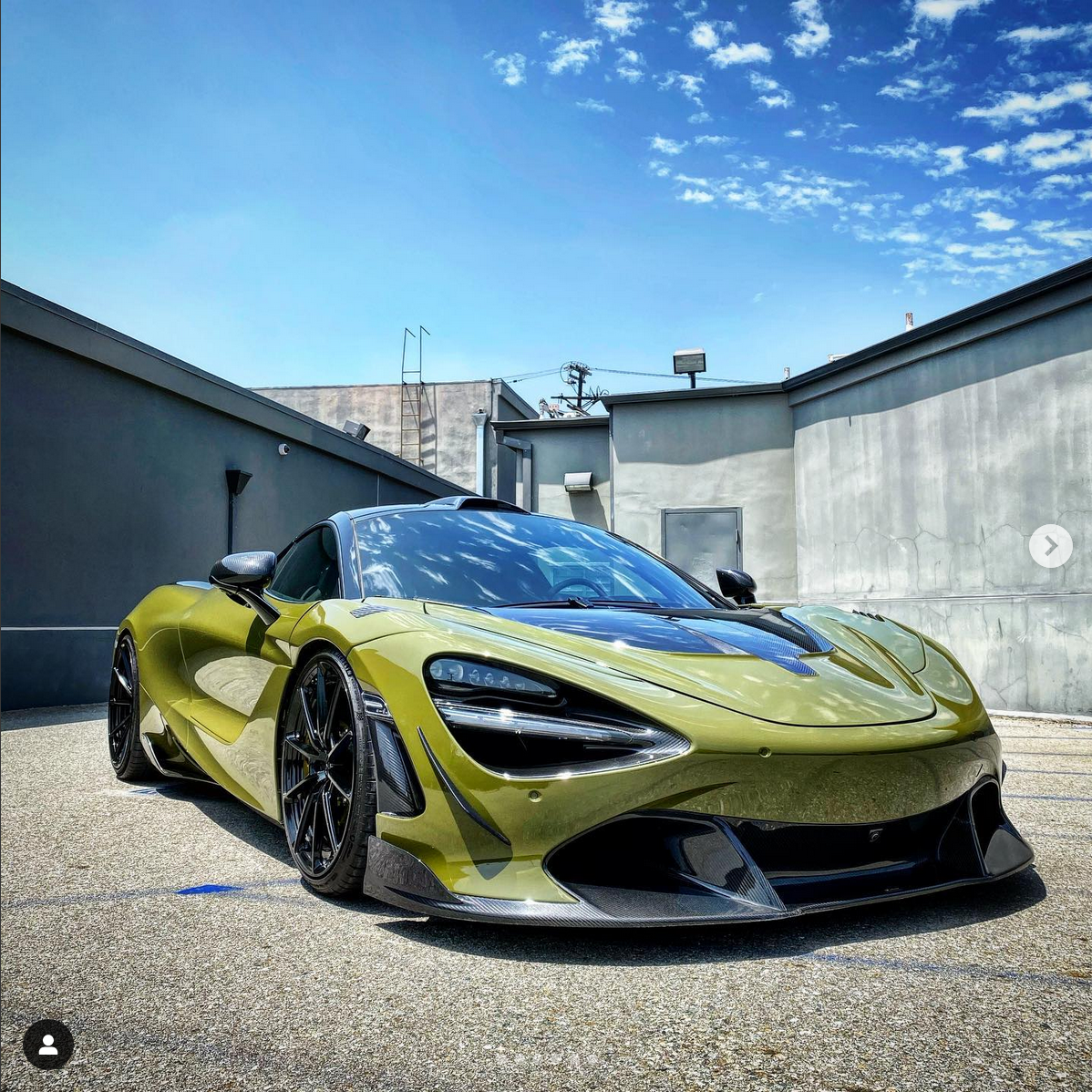 A yellow and black sports car parked in an outdoor lot with gray walls and a clear blue sky.
