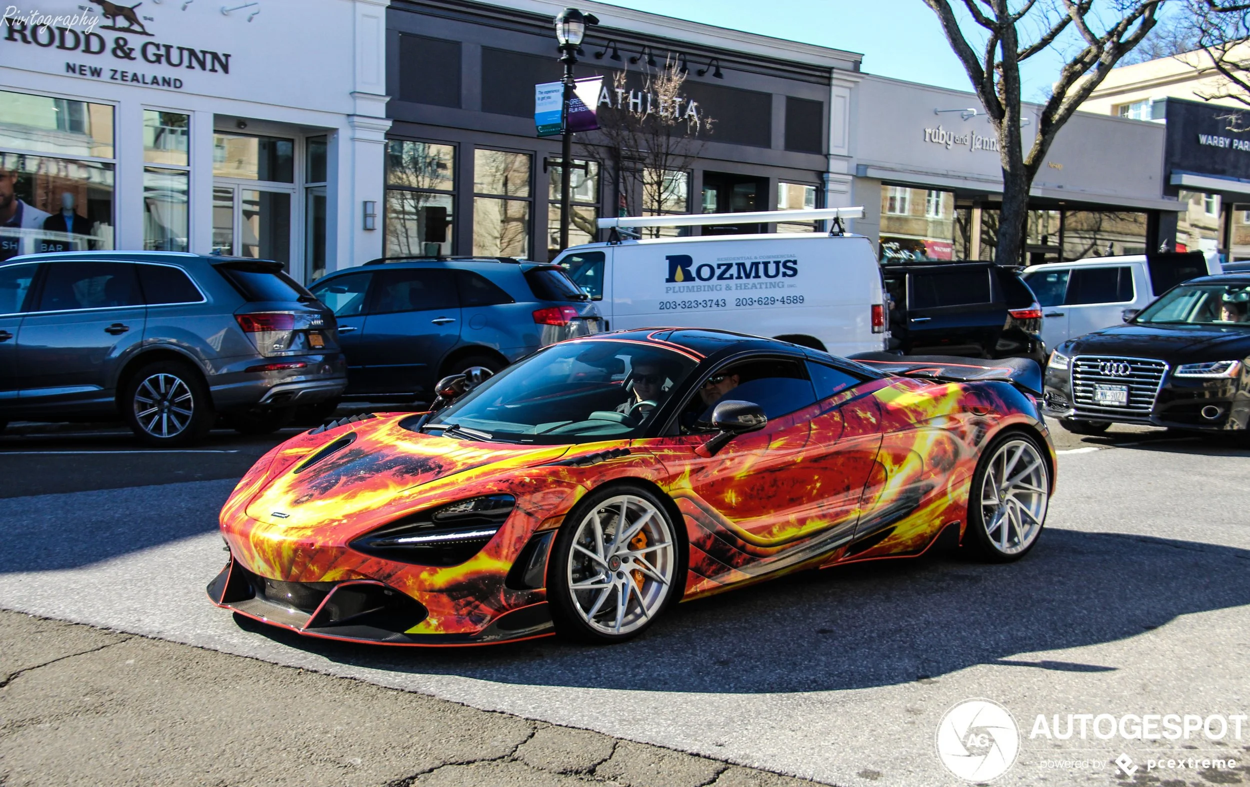 A bright red and yellow flame-themed sports car parked on a city street, with other cars in the background and storefronts along the sidewalk.