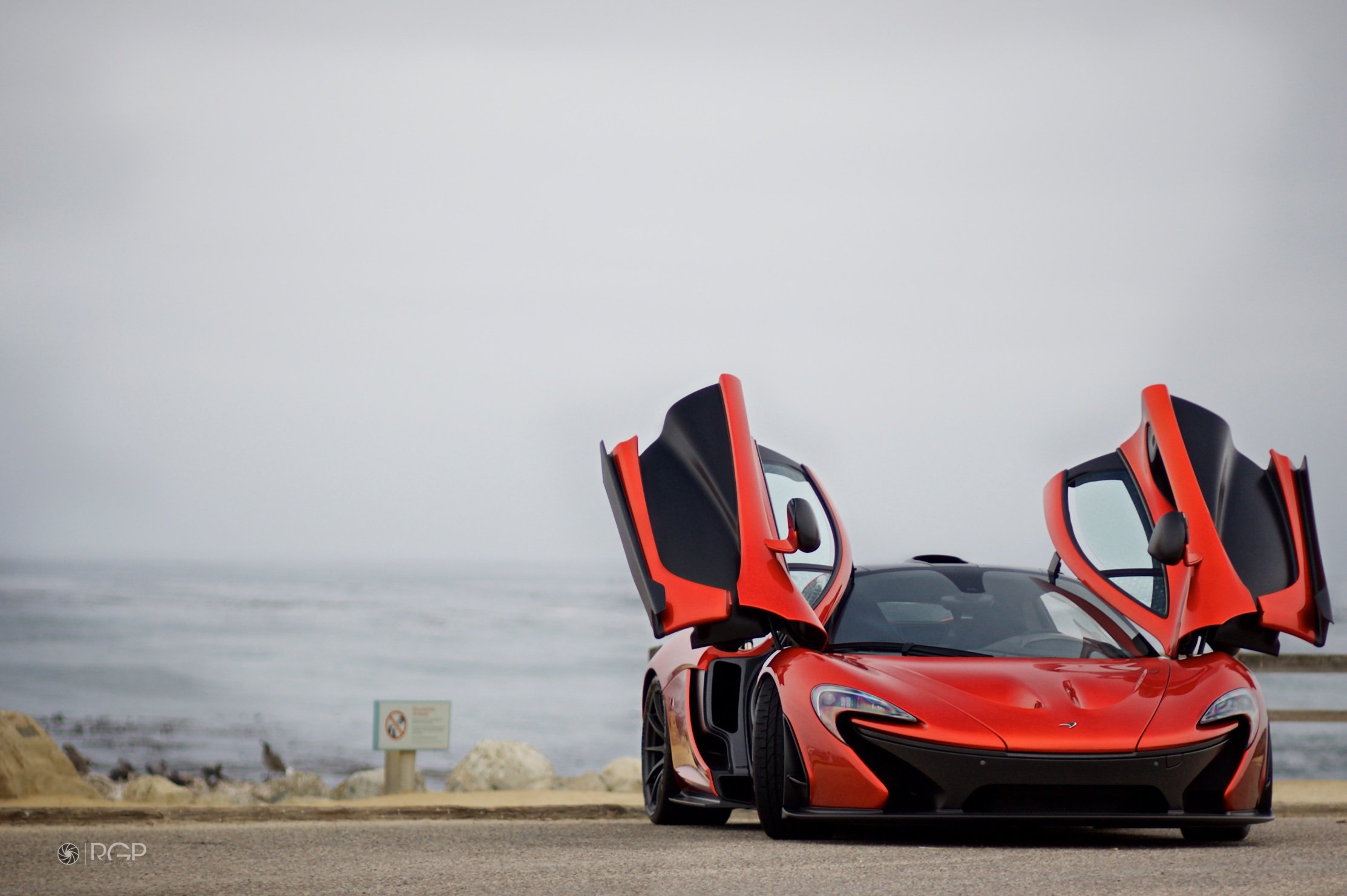 A red sports car with gull-wing doors open parked on a road near the coast, with ocean waves and a cloudy sky in the background.
