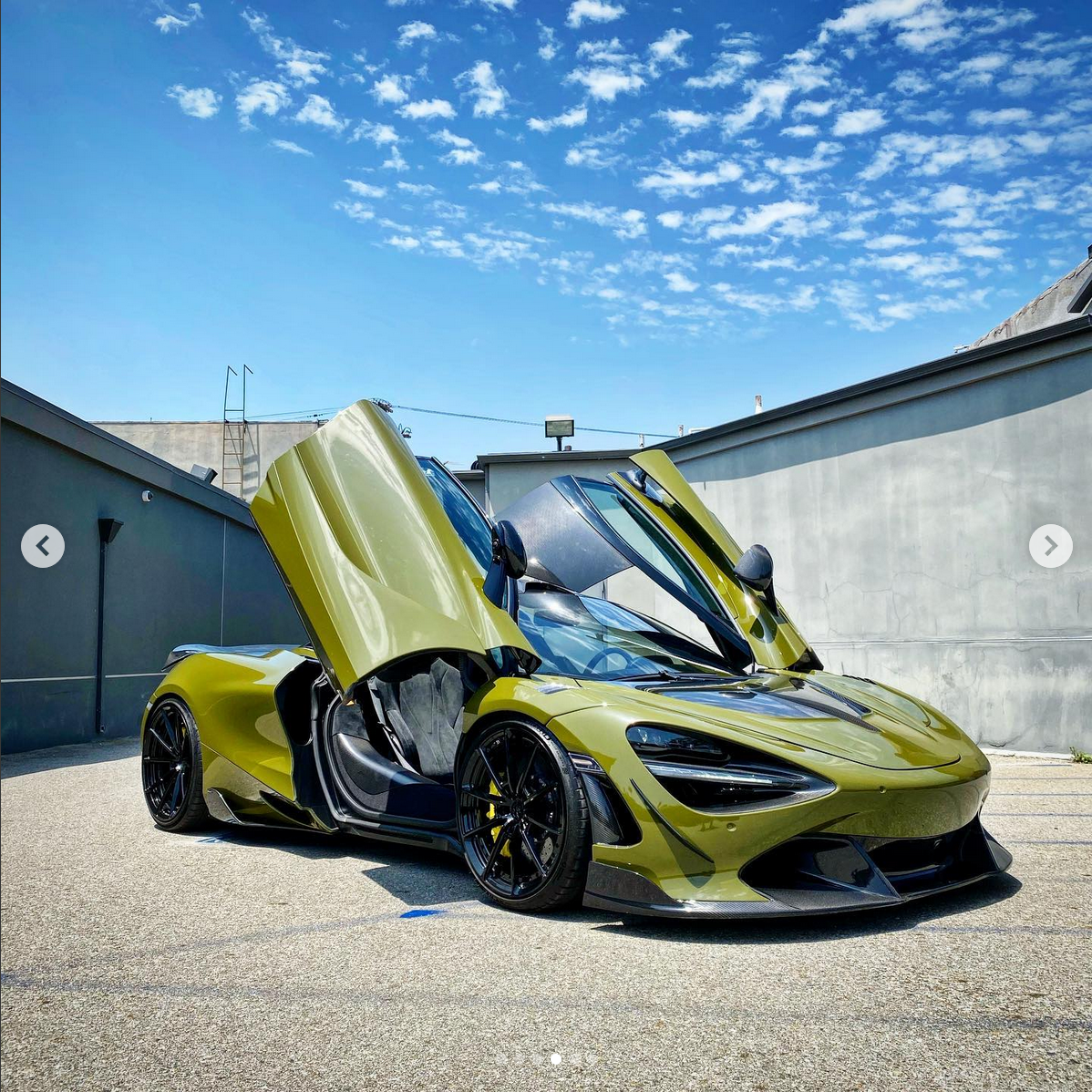 A yellow sports car with scissor doors open, parked on an outdoor surface under a blue sky with scattered clouds.