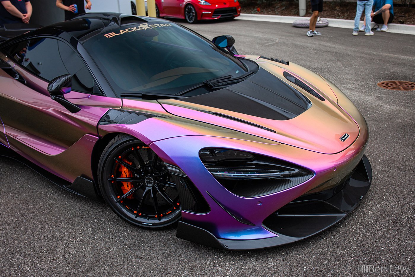 A multicolored McLaren racing car with a gradient of purple, pink, and gold is parked outdoors at a car show, with people in the background.