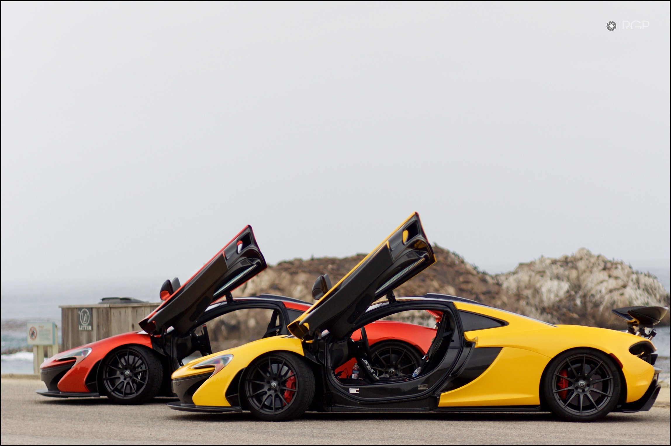 Two sports cars, one yellow and one orange, with butterfly doors open, parked near the coastline with rocky hills in the background.