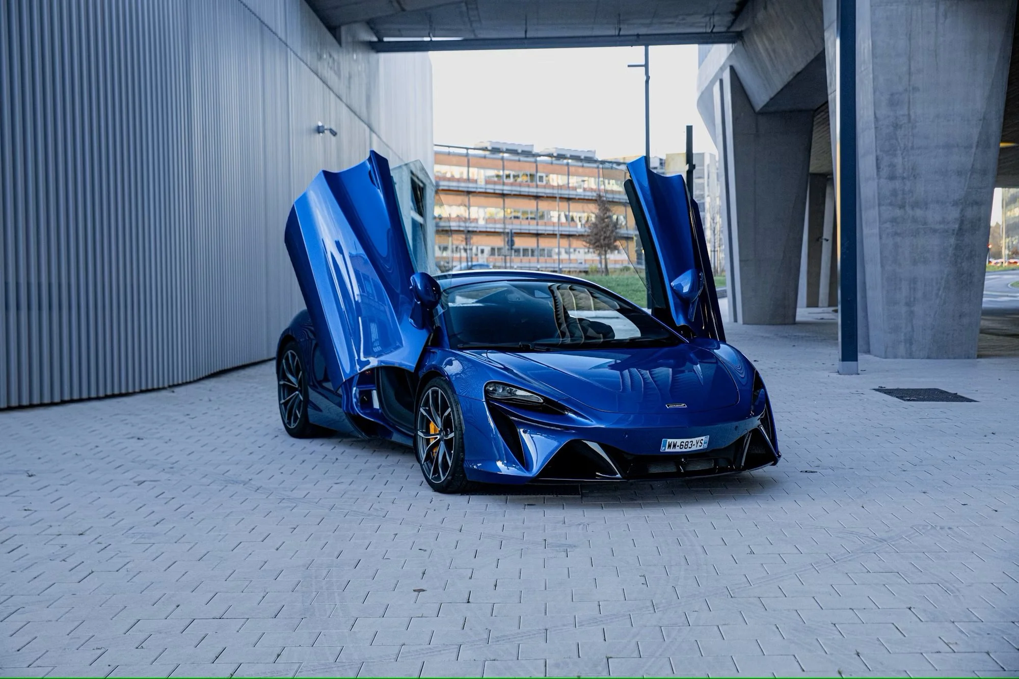 A blue luxury sports car with vertical doors open, parked on a paved area near a modern building and under an overpass.