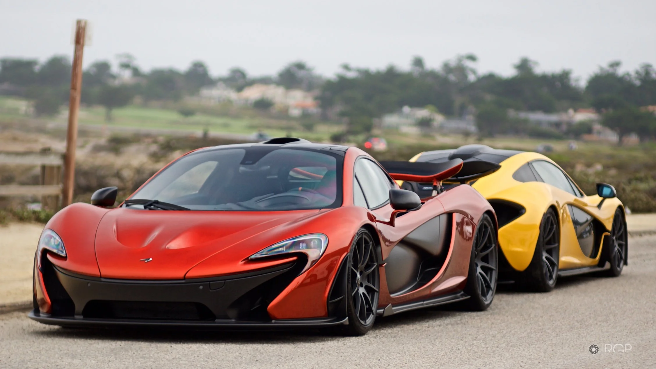 A red and black McLaren sports car parked on the side of a road with a yellow and black sports car behind it.