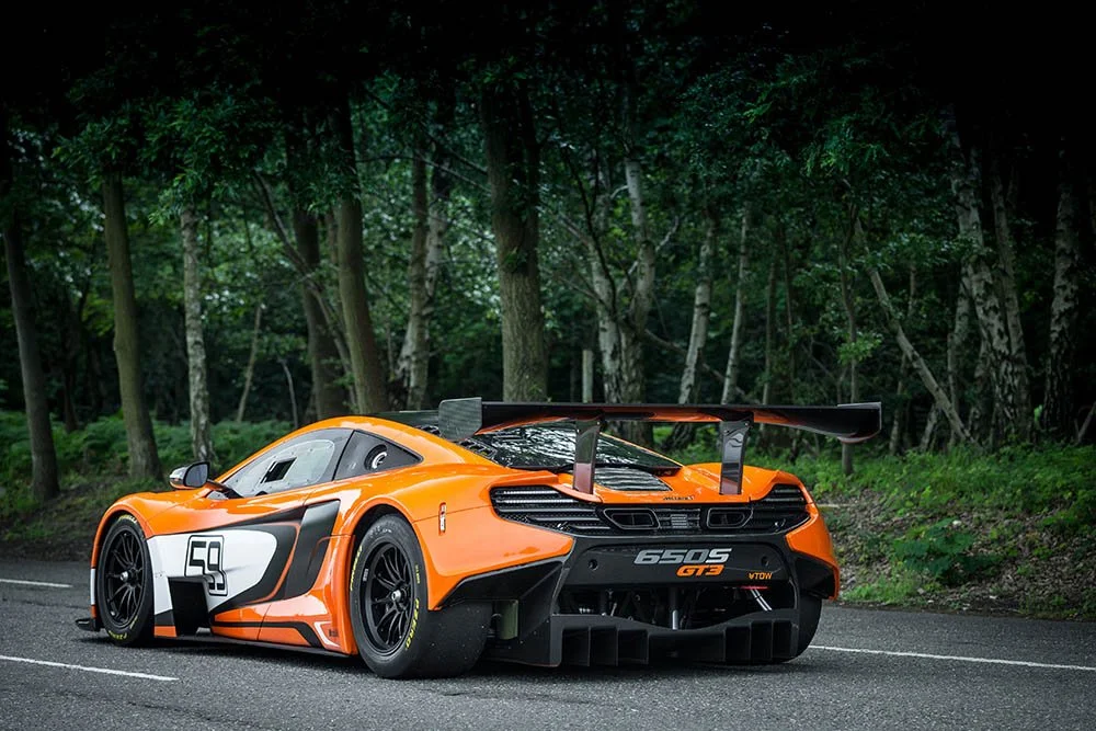 An orange racing sports car with a large rear wing on a road surrounded by trees.
