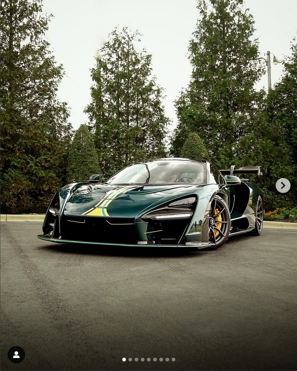 A sleek green sports car with yellow and black racing stripes parked on an asphalt road, surrounded by tall trees and greenery.