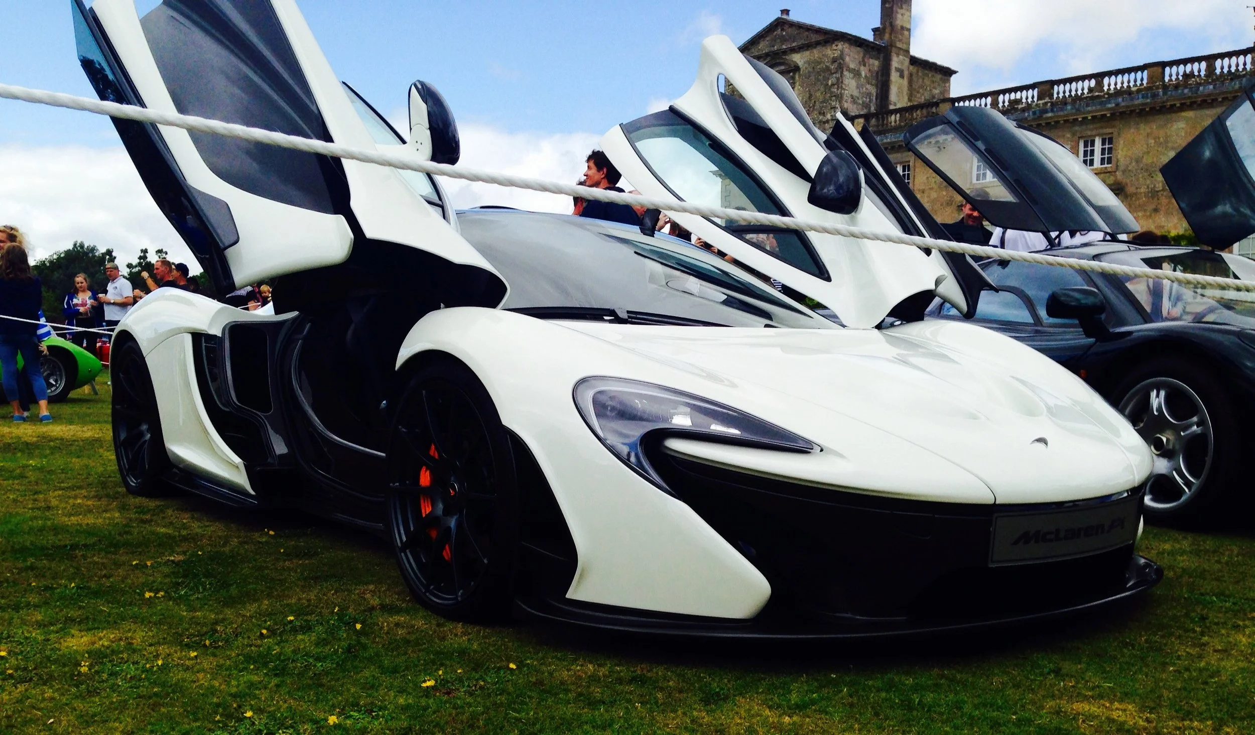 A white McLaren sports car with butterfly doors open, displayed at an outdoor car show, with people and other cars in the background.