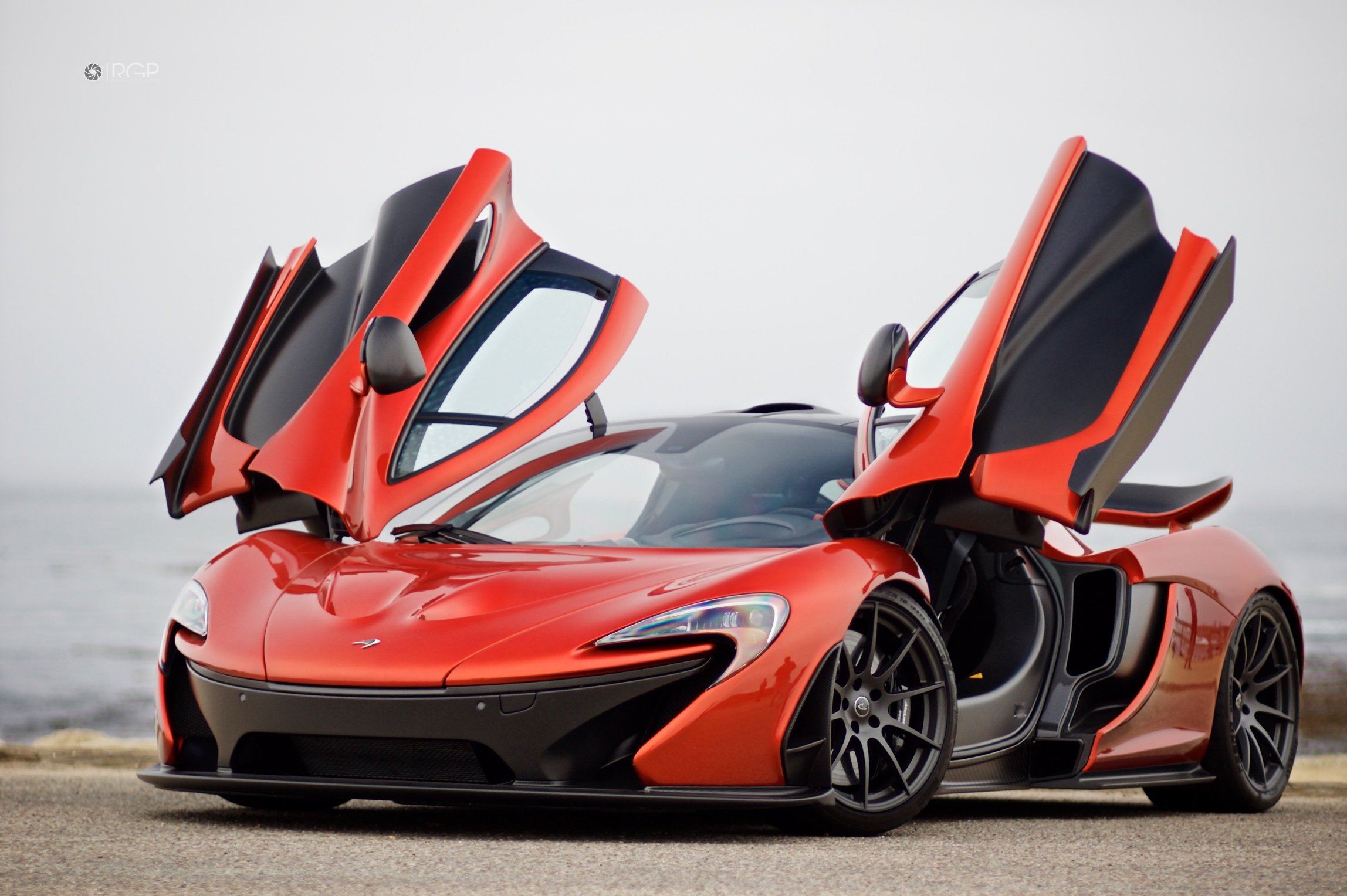 A red luxury sports car with its gull-wing doors open, parked on a road near a body of water.
