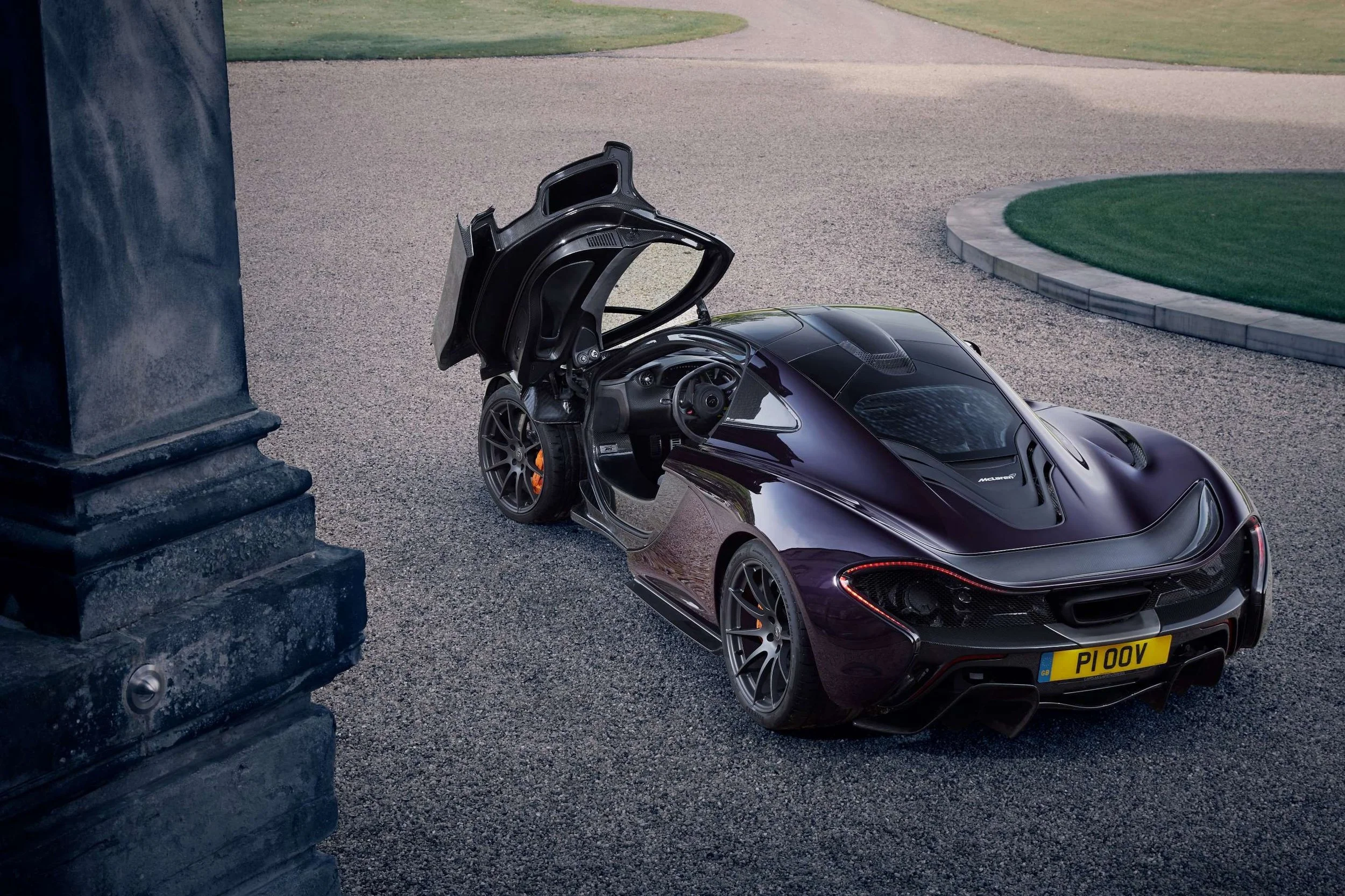 A black sports car with butterfly doors open, parked on a paved surface near a stone pillar and a grassy area.