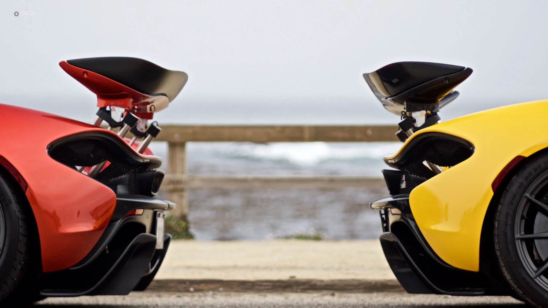 Close-up of two sports cars, one red and one yellow, parked facing each other with their rear ends visible, with a blurry seaside background and a wooden fence.
