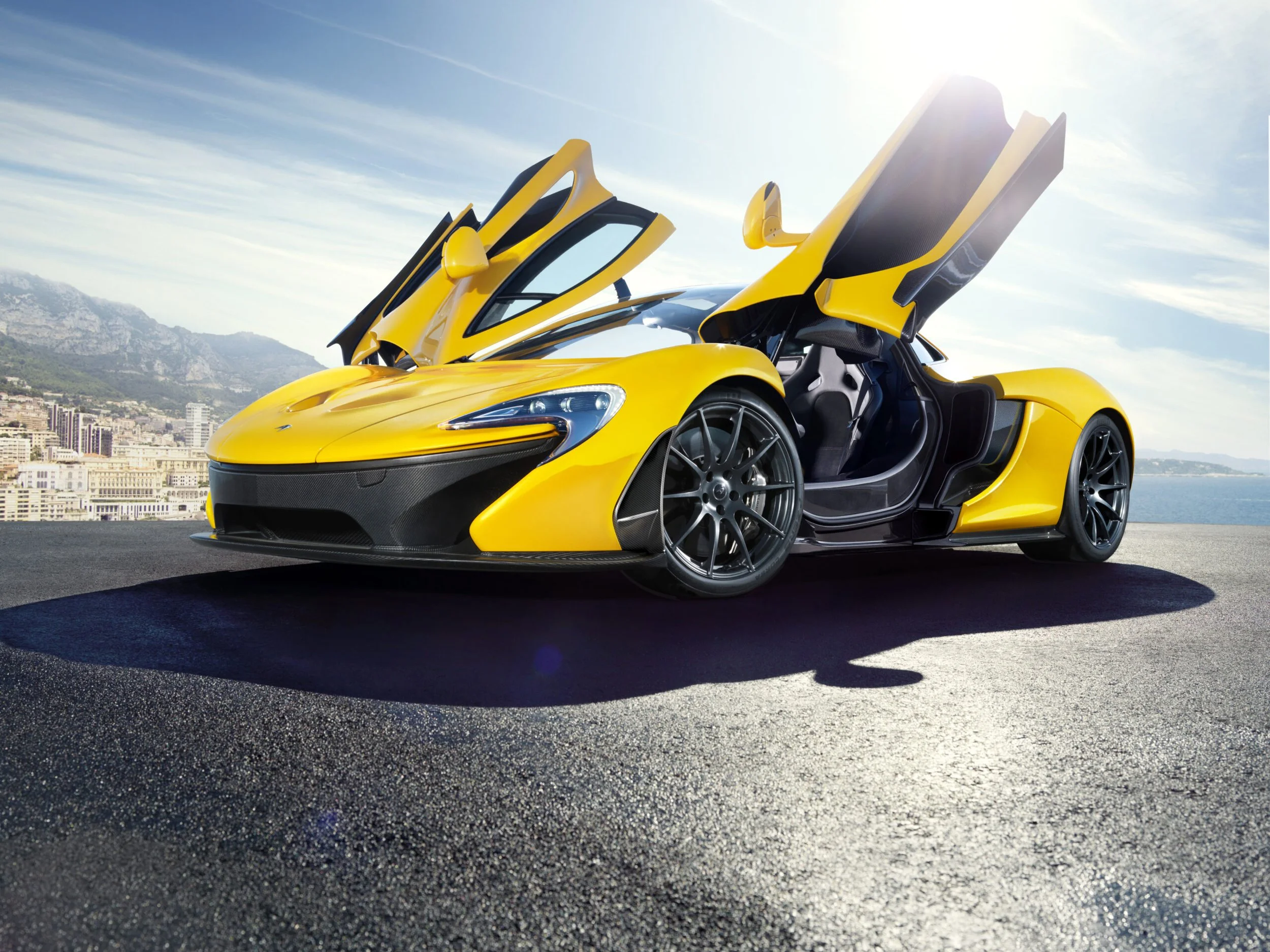 A yellow luxury sports car with upward-opening doors parked on a rooftop with a city and mountains in the background.