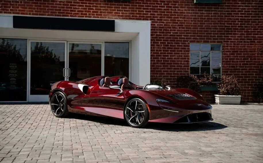 Red sports car with open cockpit parked on a brick-paved area in front of a building with large glass doors and red brick walls.