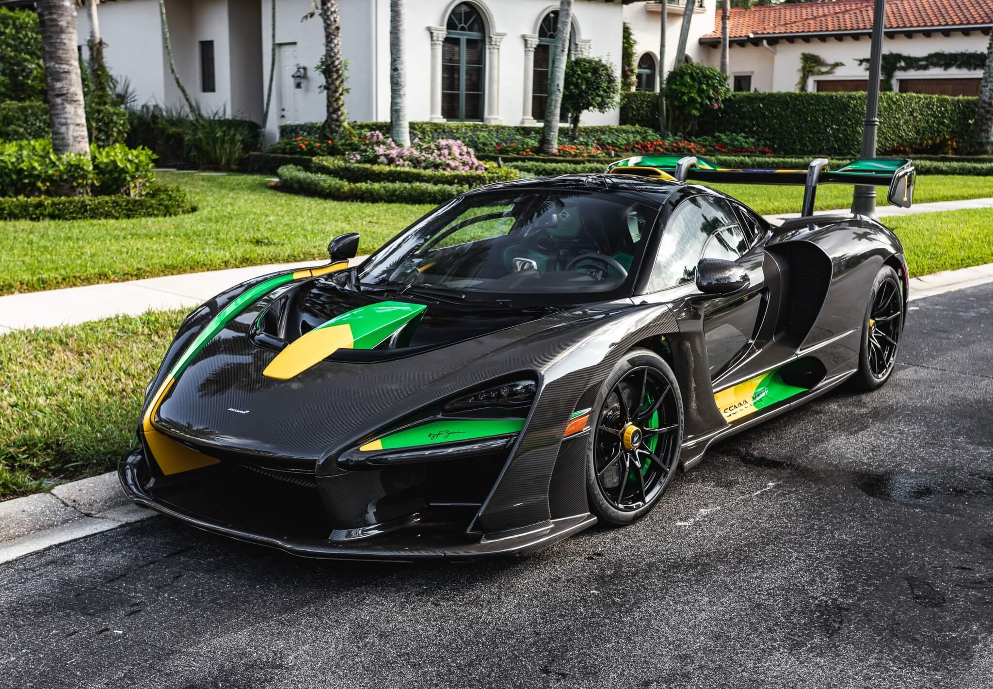 Black sports car with green and yellow racing decals parked on a residential street in front of a white house with a well-kept lawn and landscaped garden.