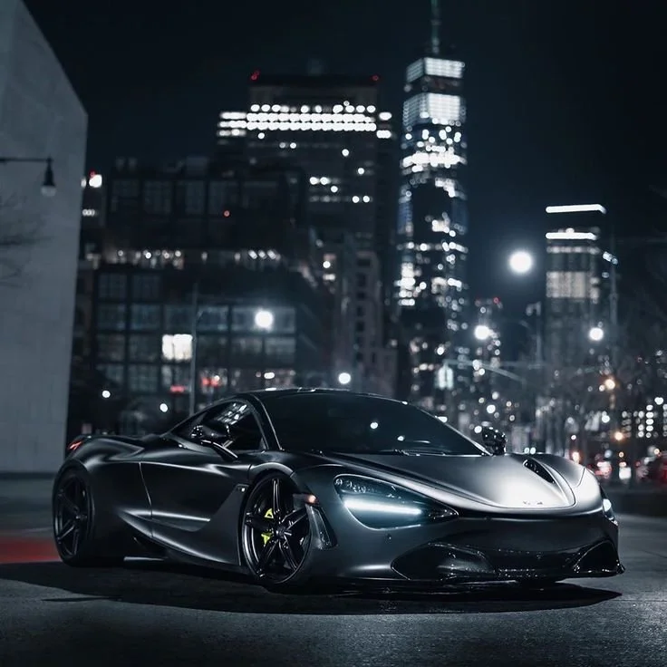 A sleek black sports car parked on a city street at night with illuminated skyscrapers in the background.