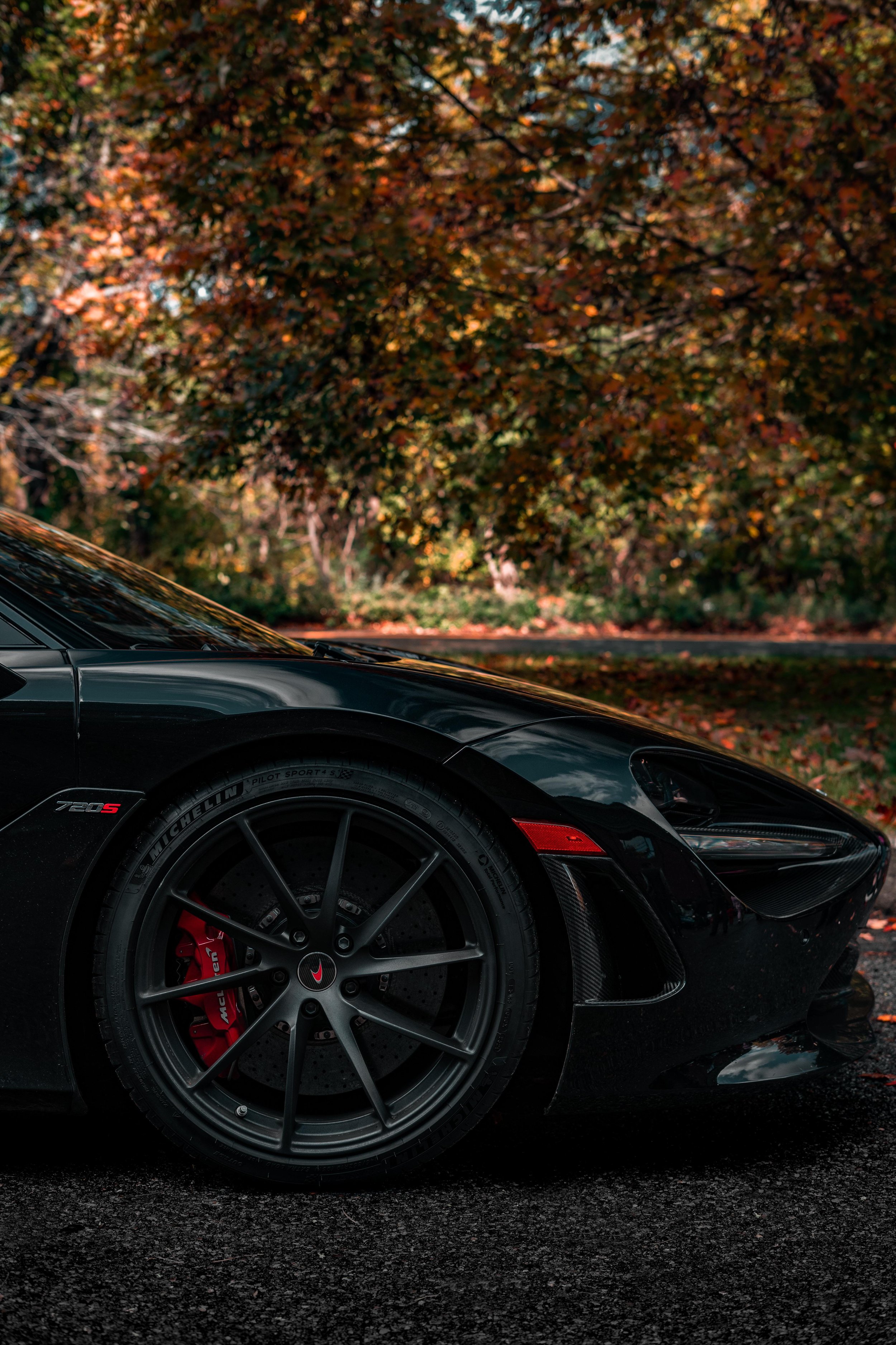 Close-up of the front part of a black sports car with red brake calipers, parked on a dark road with autumn-colored trees in the background.