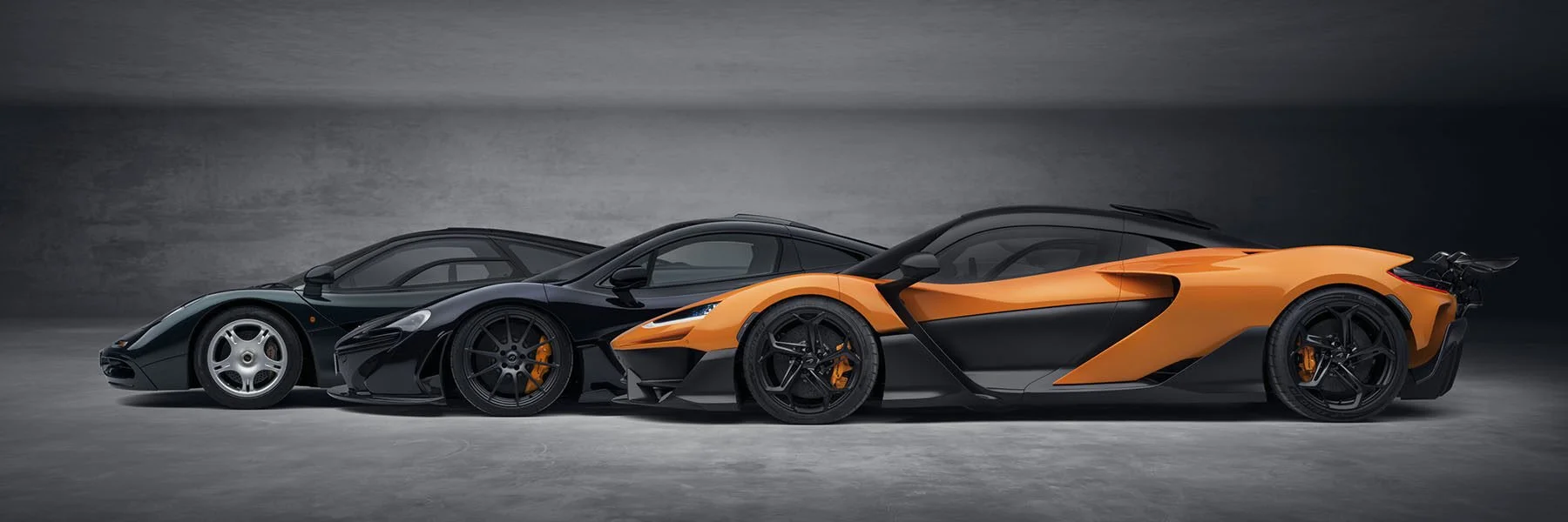 Side view of three luxury sports cars, with the first being black, the second black, and the third orange, parked in a row in front of a gray background.