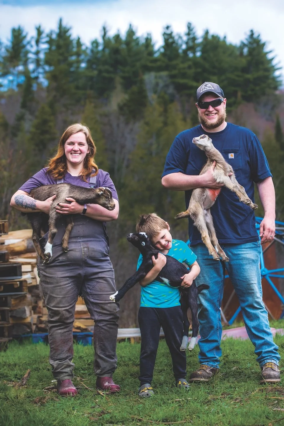 A family of farmers—a woman, small child, and man—hold young goats on a farm with trees in the background.