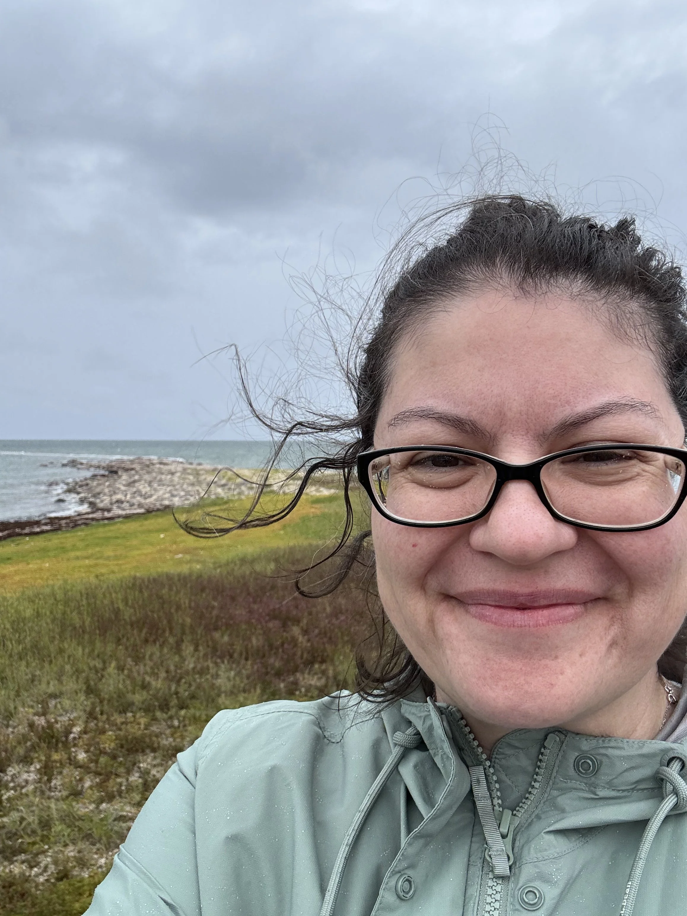 A woman with glasses smiling outdoors on a cloudy day, wearing a light green jacket, with a grassy area and rocky shoreline in the background.
