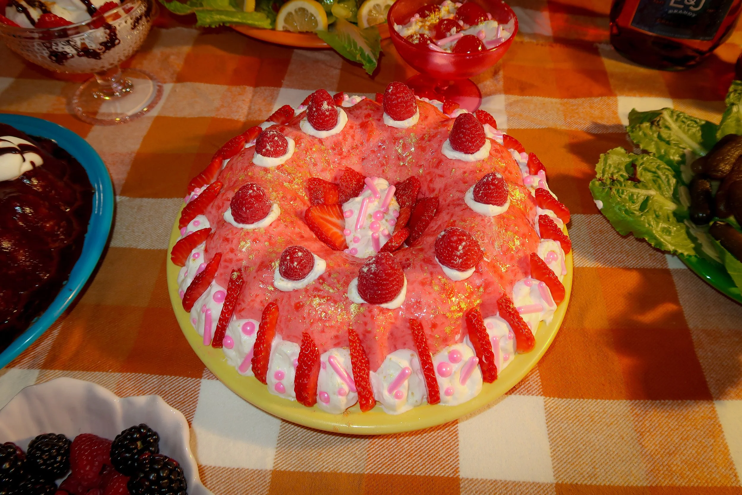 A pink frosted cake decorated with fresh raspberries, strawberries, and colorful sprinkles on a yellow platter, surrounded by bowls of berries, salad, and other desserts on a checkered tablecloth.