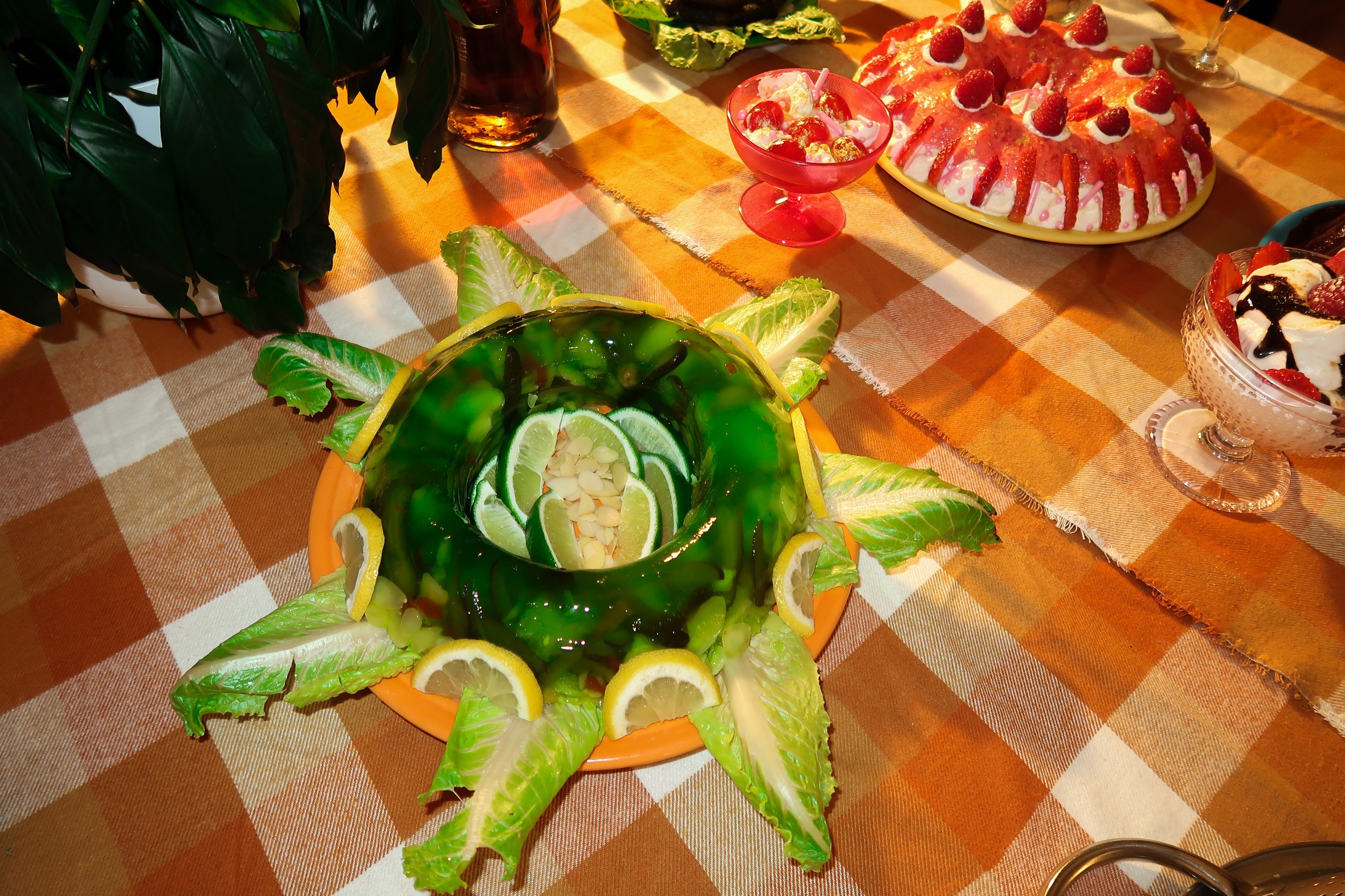 Colorful party table with a large strawberry and cream cake decorated with strawberries, a glass bowl of dessert with fruit toppings, and a green jelly mold with lemon and lime slices, all on a checkered orange and white tablecloth.