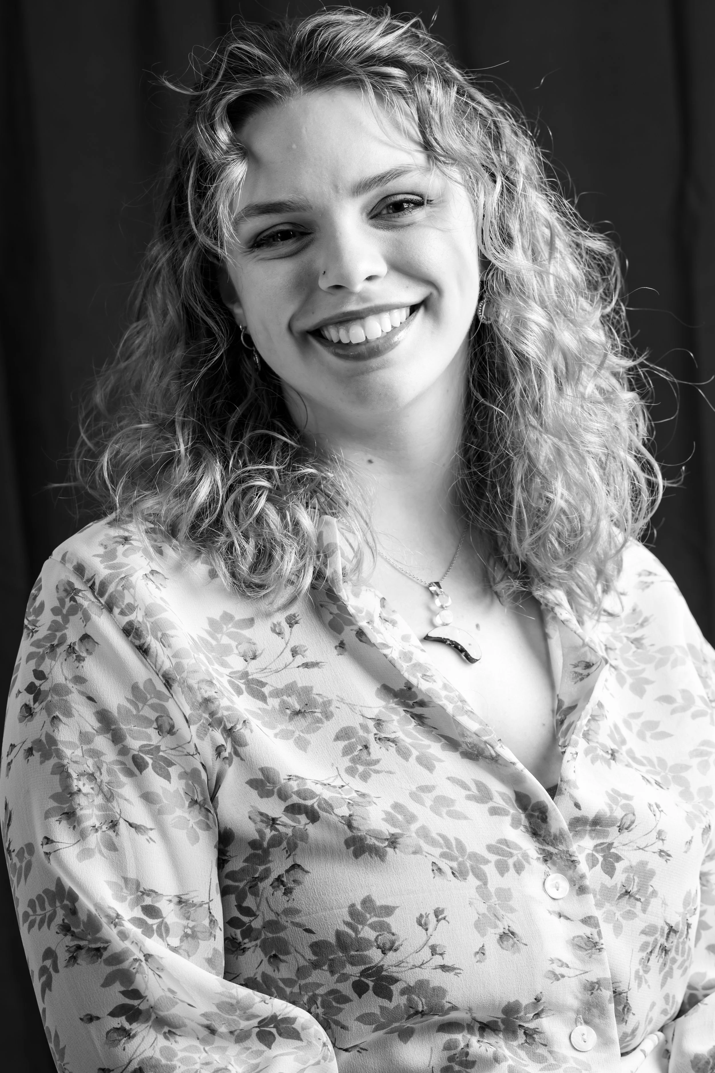 Black and white portrait of a woman with curly hair smiling and looking at the camera, wearing a floral patterned shirt and jewelry.