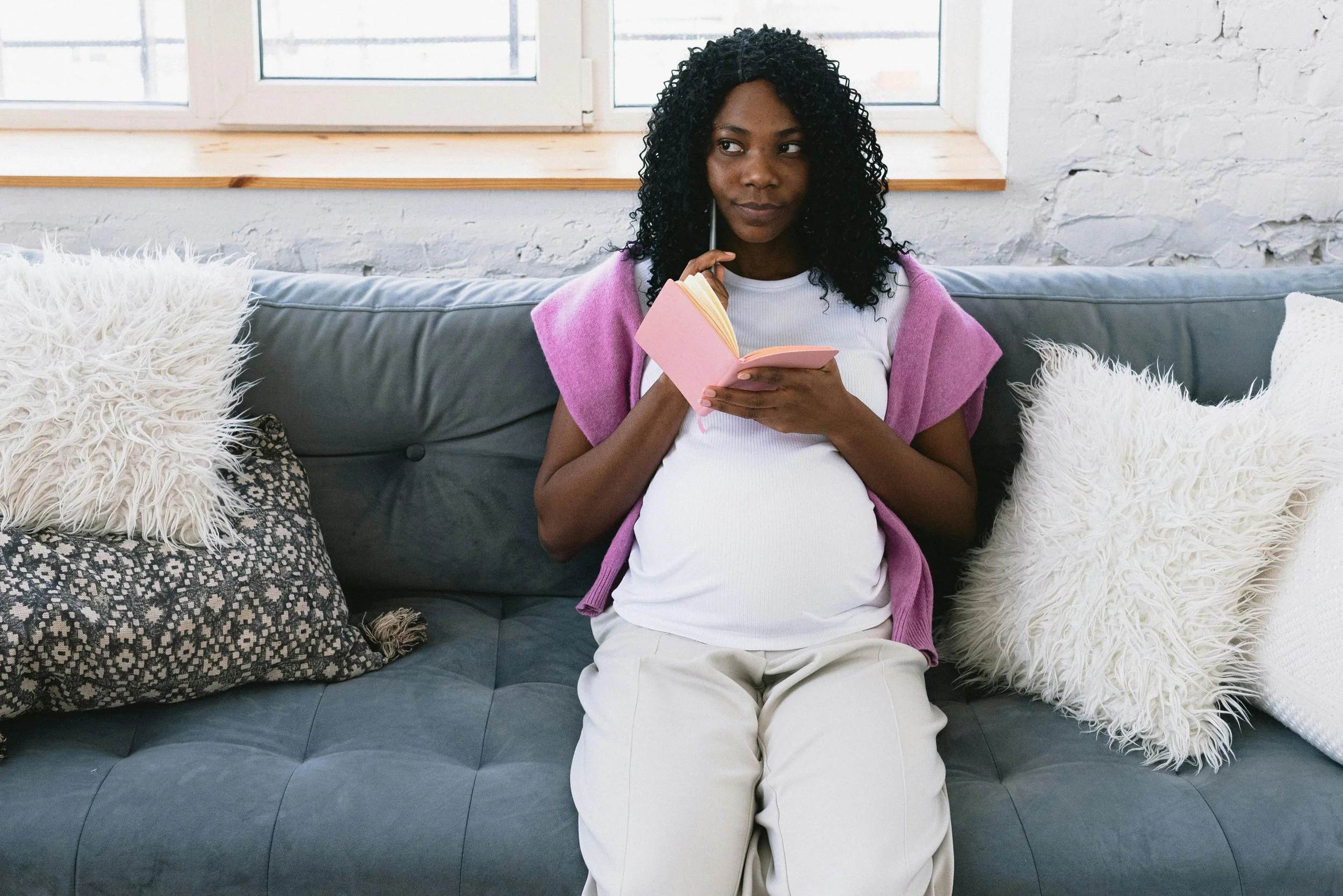 Pregnant woman sitting on a gray couch with white and black pillows, holding a pink notebook, wearing a white shirt and beige pants, creating her birth prefernces plan.