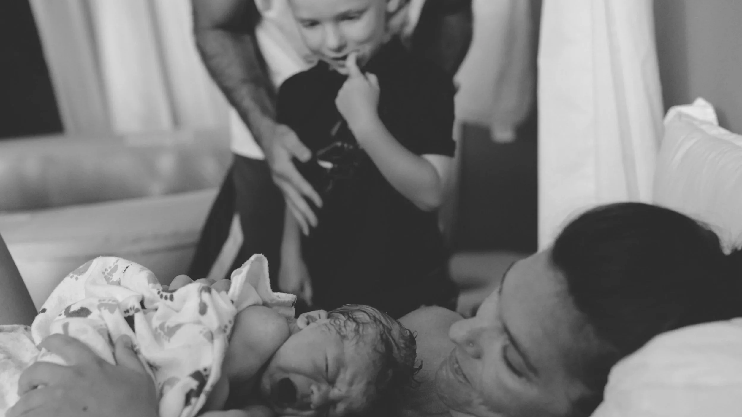 Black and white photo of a woman lying in a hospital bed holding a newborn baby. A young boy stands next to her, looking at the baby and woman with curiosity.