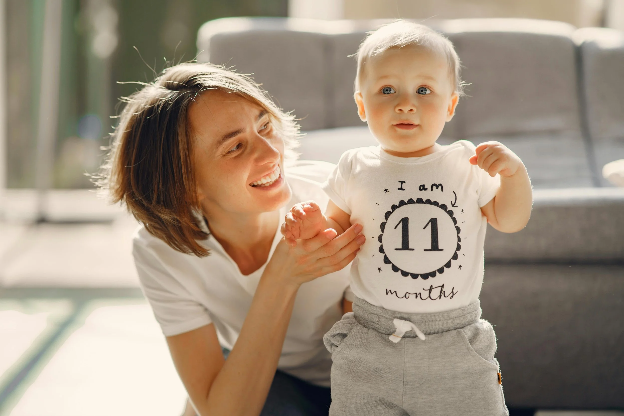 A woman with short brown hair smiling and holding a young boy who is standing. The boy has short blonde hair and is wearing a white T-shirt with the text 'I am 11 months' and gray pants.