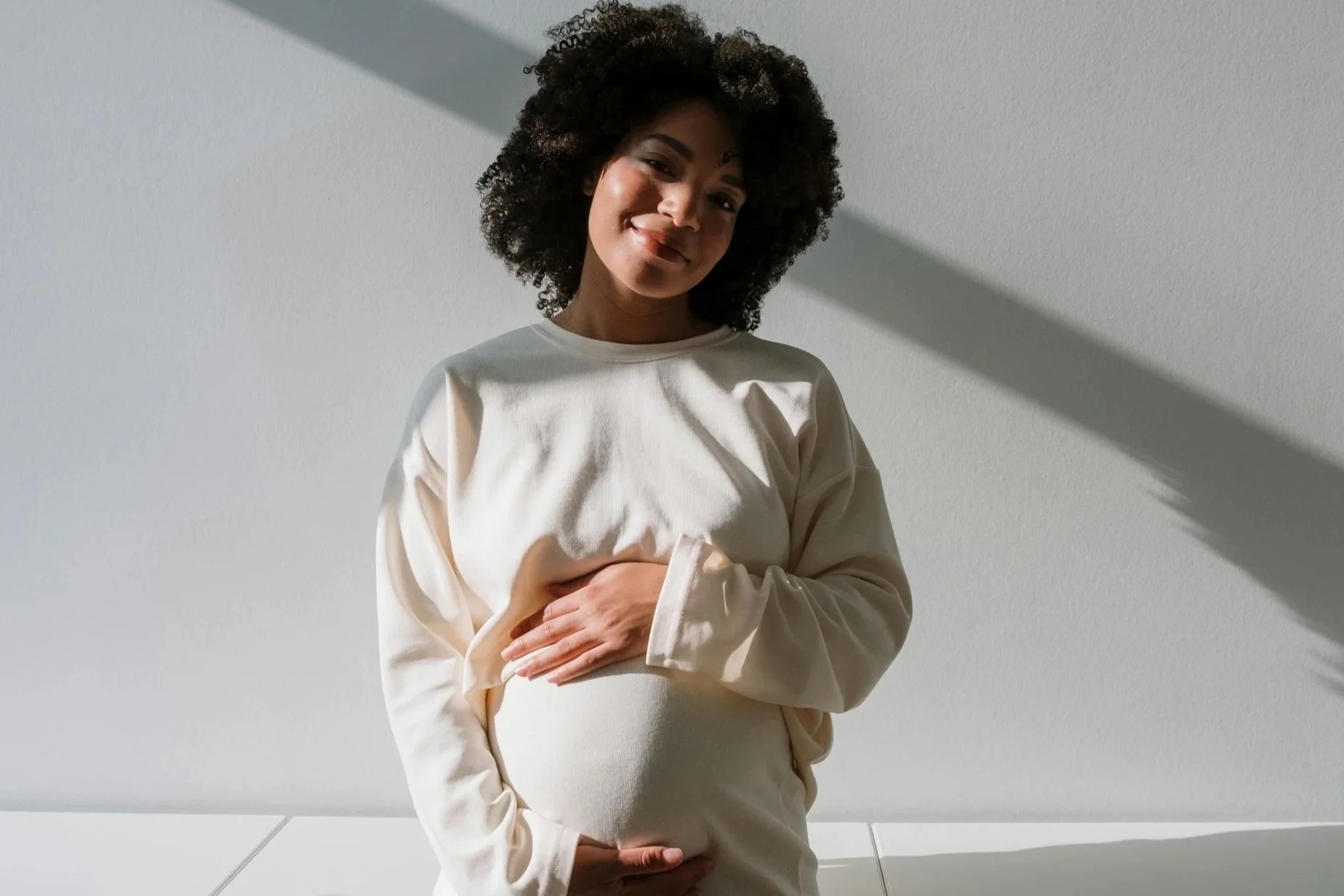 A pregnant woman with curly hair smiling and gently touching her belly, standing against a white wall with sunlight casting a shadow.