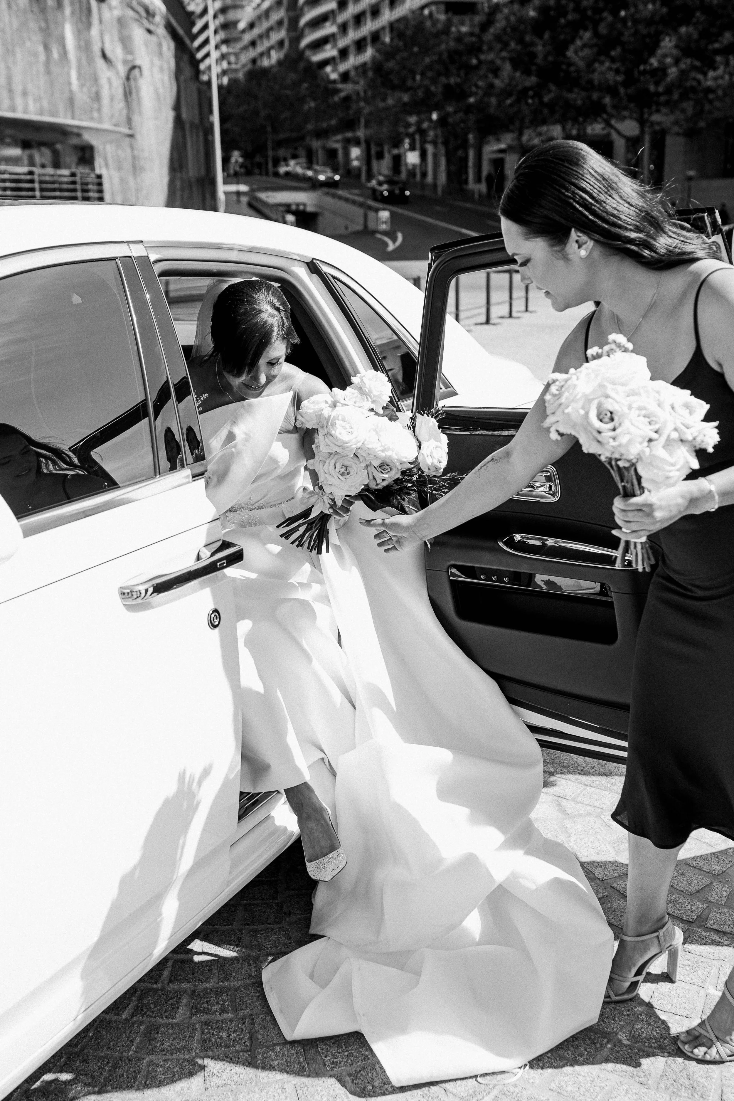 A bride getting out of a luxury car with the help of a bridesmaid, holding bouquets of flowers.