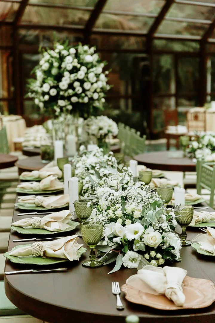 Elegant dining table with white floral centerpieces, green glasses, and neatly folded napkins inside a greenhouse-like setting.