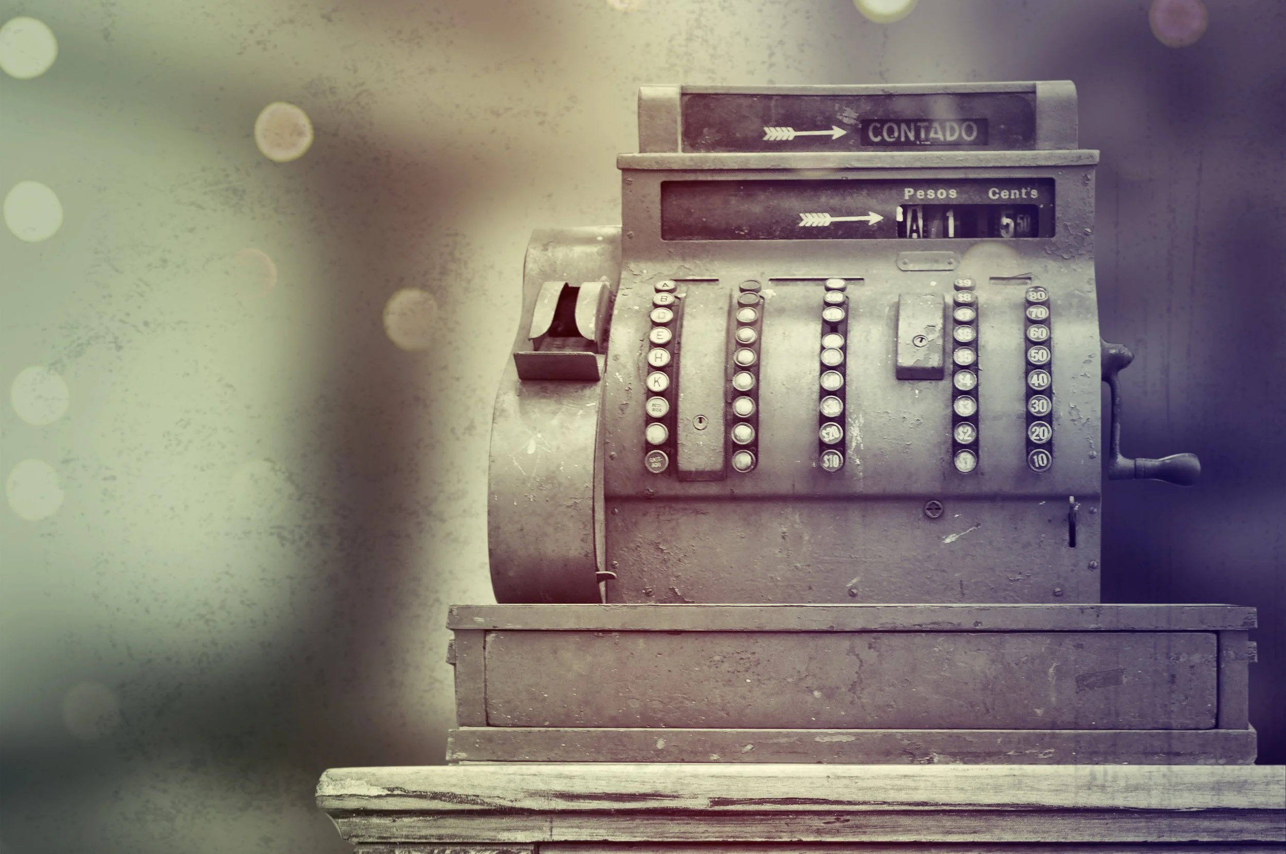 An old metallic cash register with buttons, a small display at the top showing currency, and a handle on the right side, sitting on a wooden table. The background is blurred with bokeh lights.