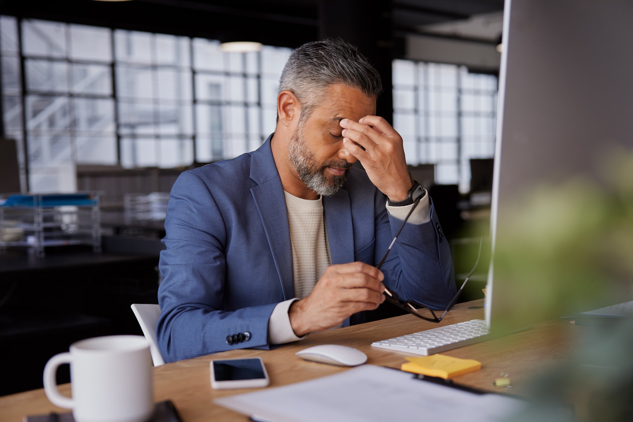 A middle-aged man with gray hair and a beard, dressed in a blue blazer, sitting at a desk in an office, holding his head with one hand and his glasses with the other, appearing stressed or frustrated.